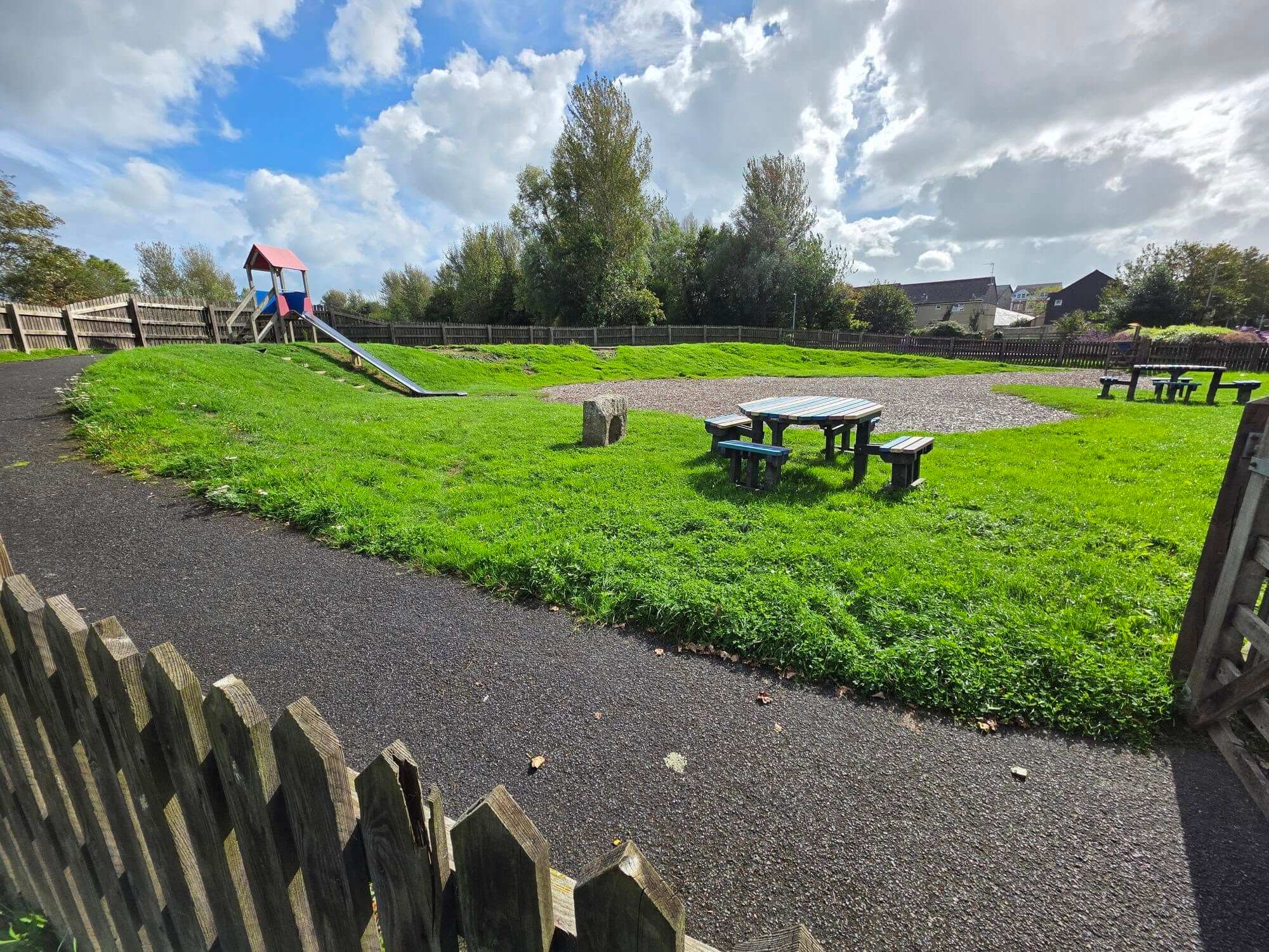 Path and picnic tables at Jubilee Play Area in Wadebridge, with the slide on the bank behind