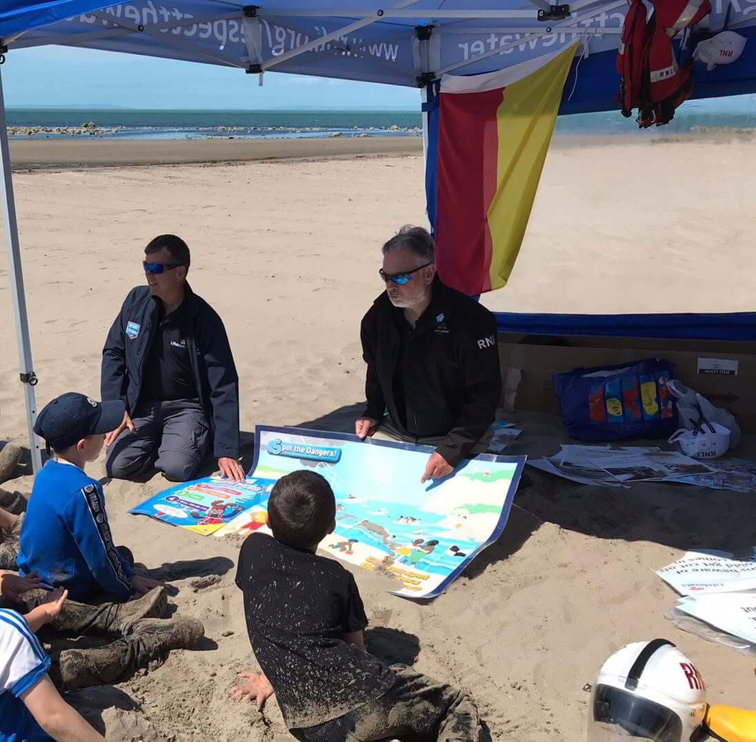 RNLI volunteers showing children a Spot the Dangers water safety poster on a beach