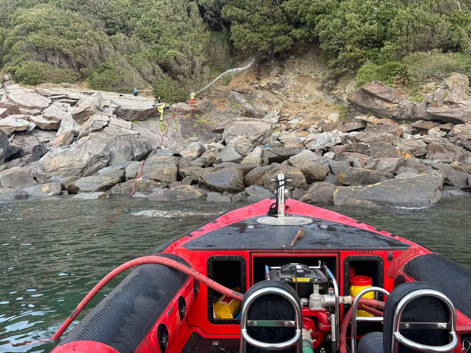 Falmouth fire boat off the rocky shore as firefighters spray water onto a wooded slope near Mawnan