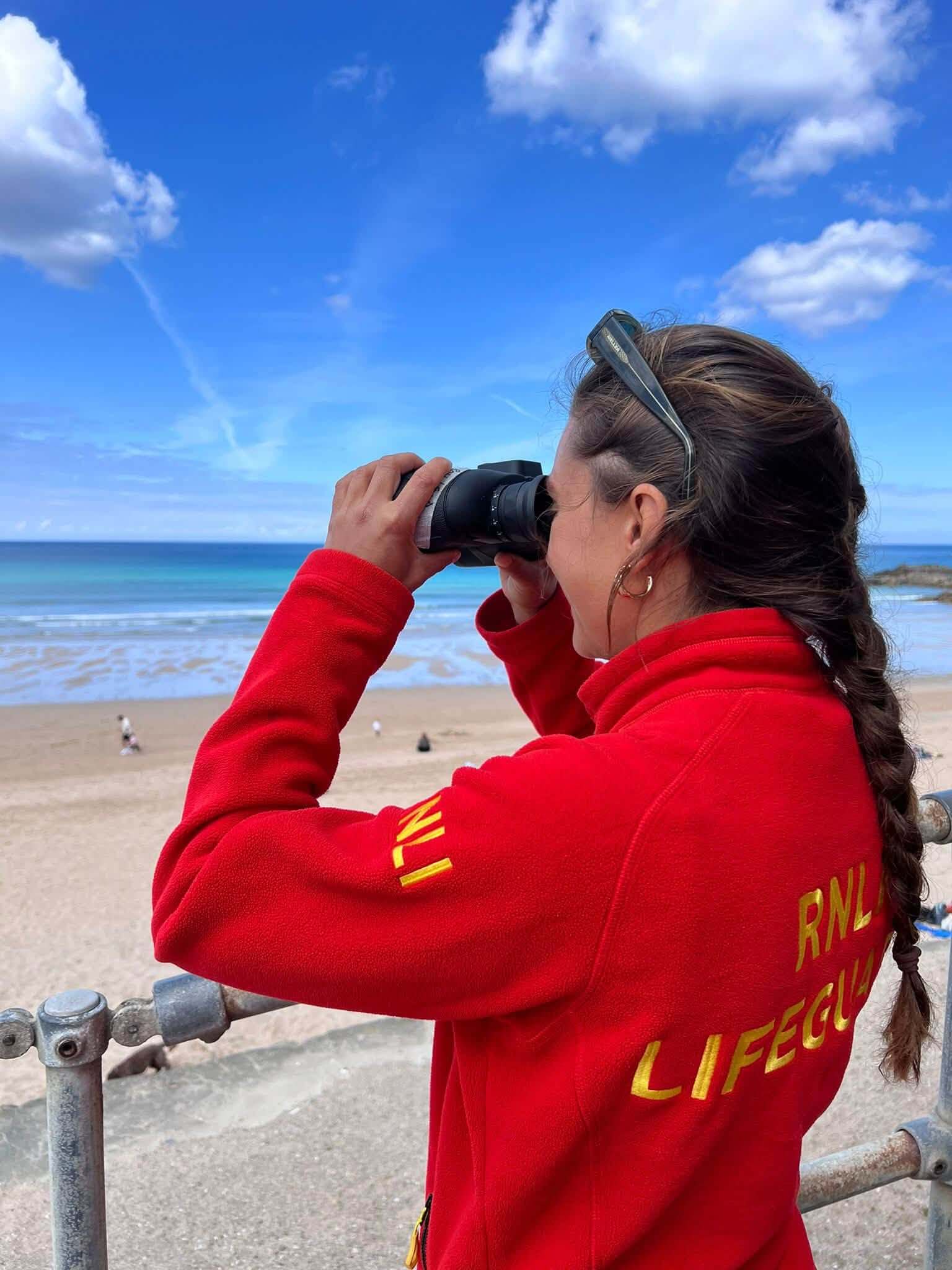 RNLI lifeguard scanning the sea through binoculars above a Newquay beach