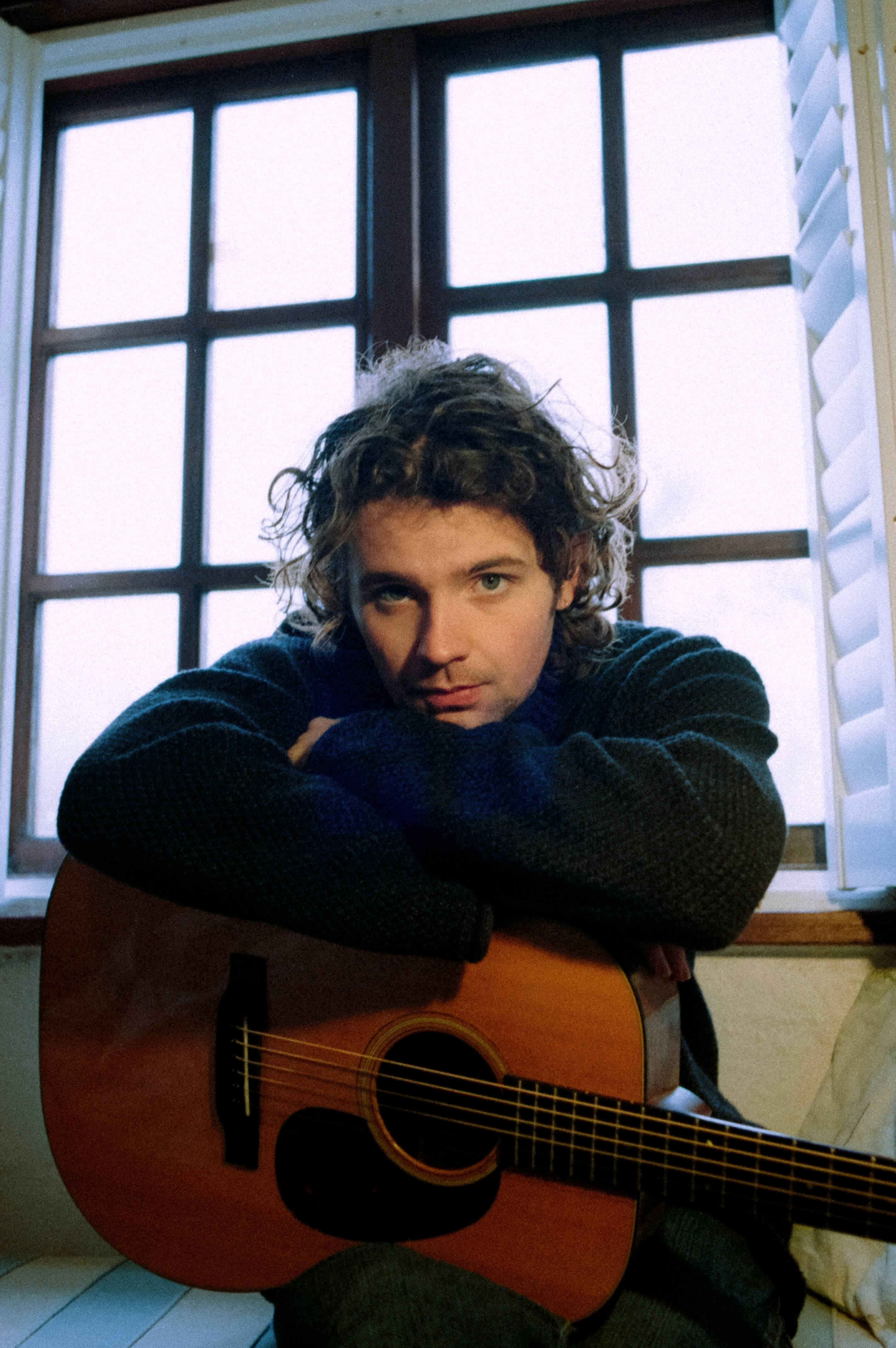 Charlie Noordewier leaning on his acoustic guitar by a window