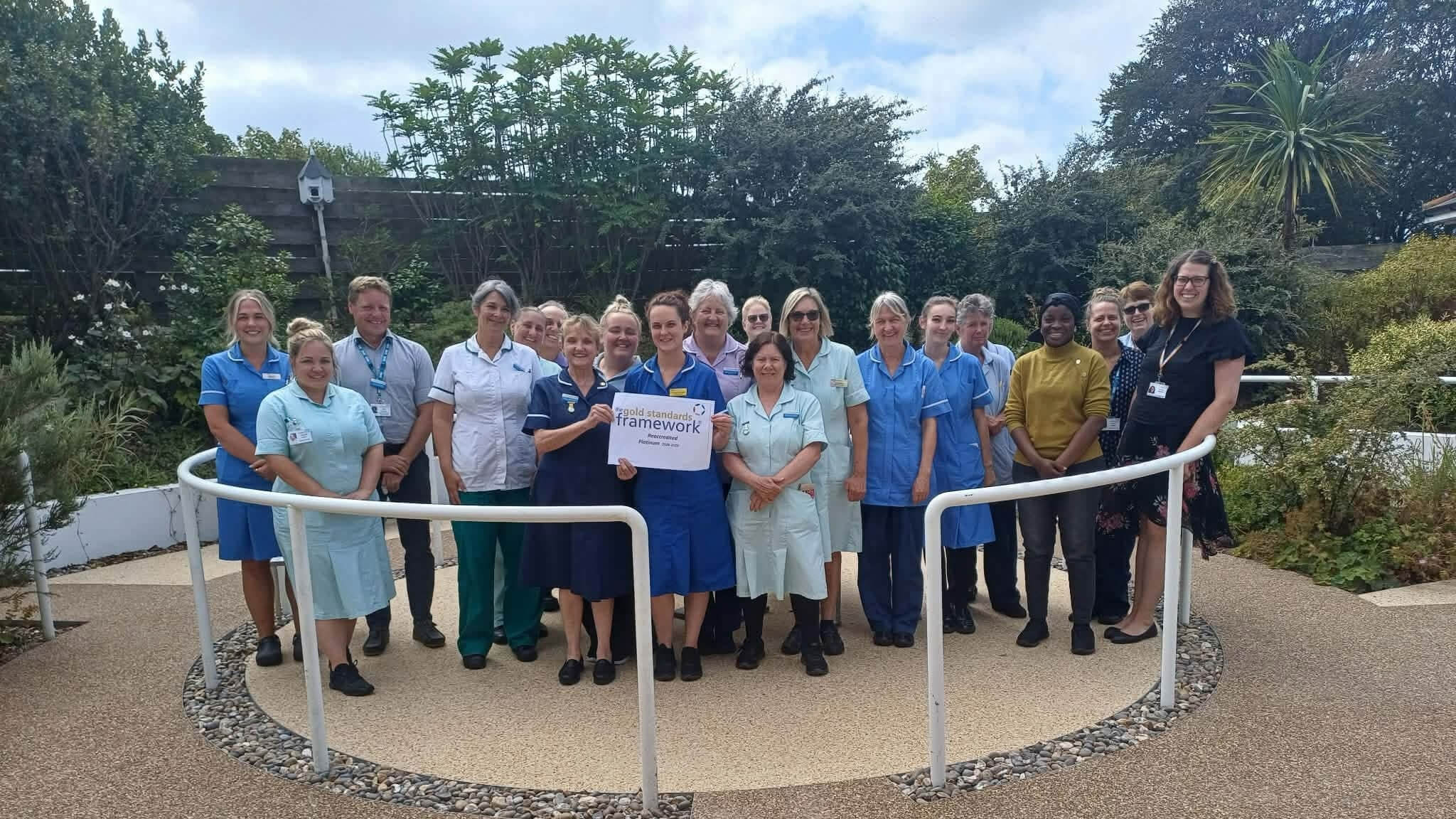 Helston Community Hospital staff holding a Gold Standards Framework platinum reaccreditation sign in the hospital garden