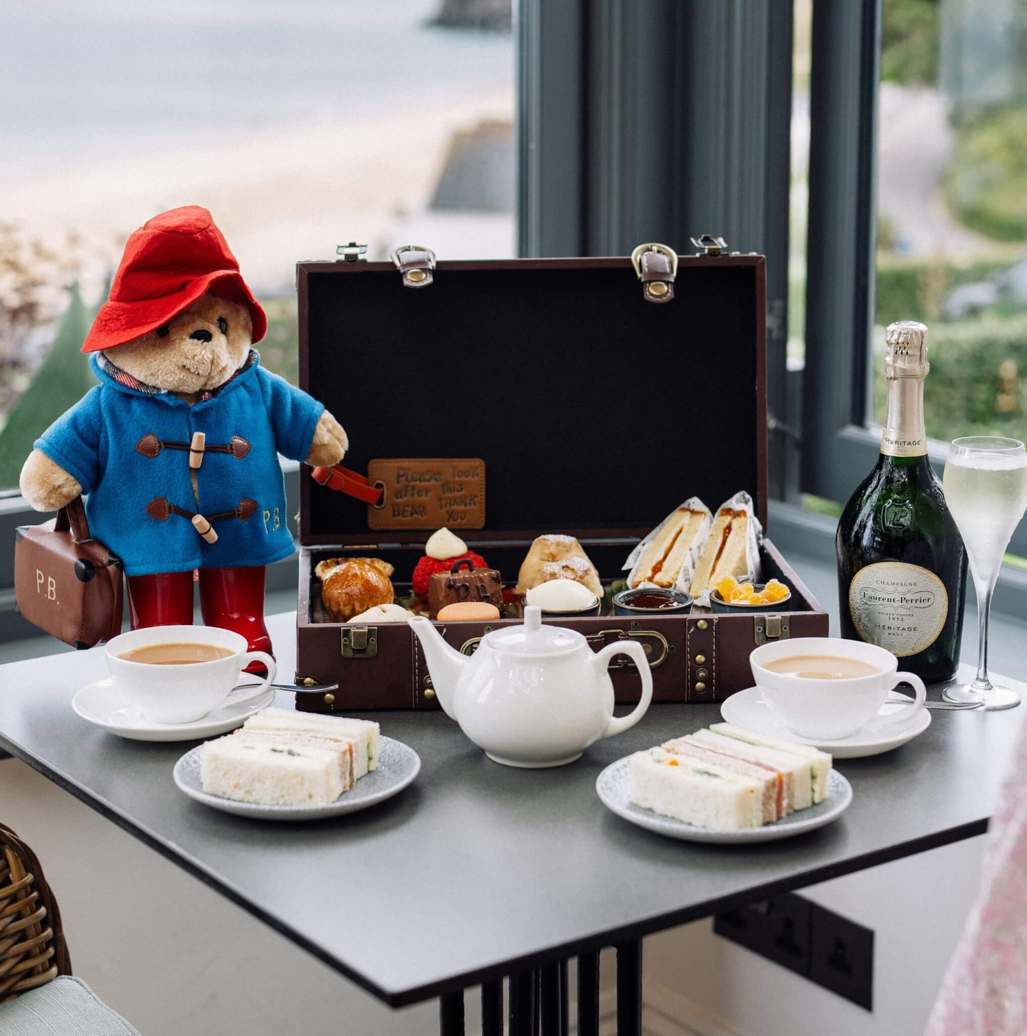 Paddington Bear beside an open suitcase of afternoon tea treats, with tea, sandwiches and Champagne by the window at Carbis Bay
