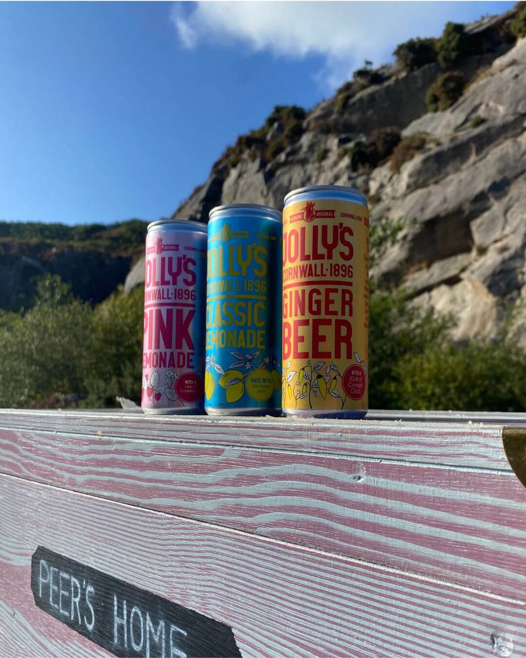 Cans of Jolly's Pink Lemonade, Classic Lemonade and Ginger Beer on a painted wooden set marked Peer's Home, in front of a rocky cliff