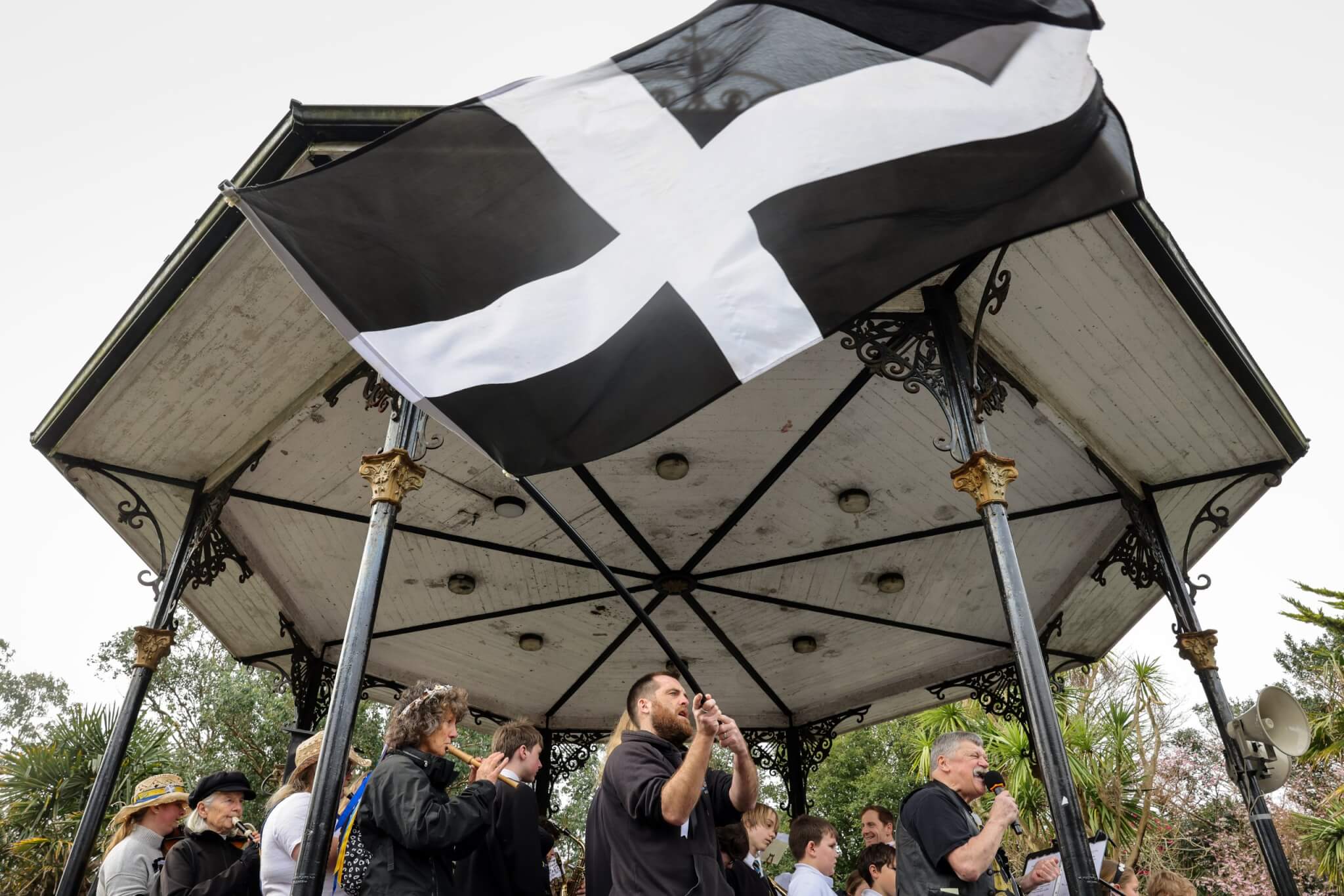 A Cornish flag flies beneath a bandstand roof as musicians play and sing