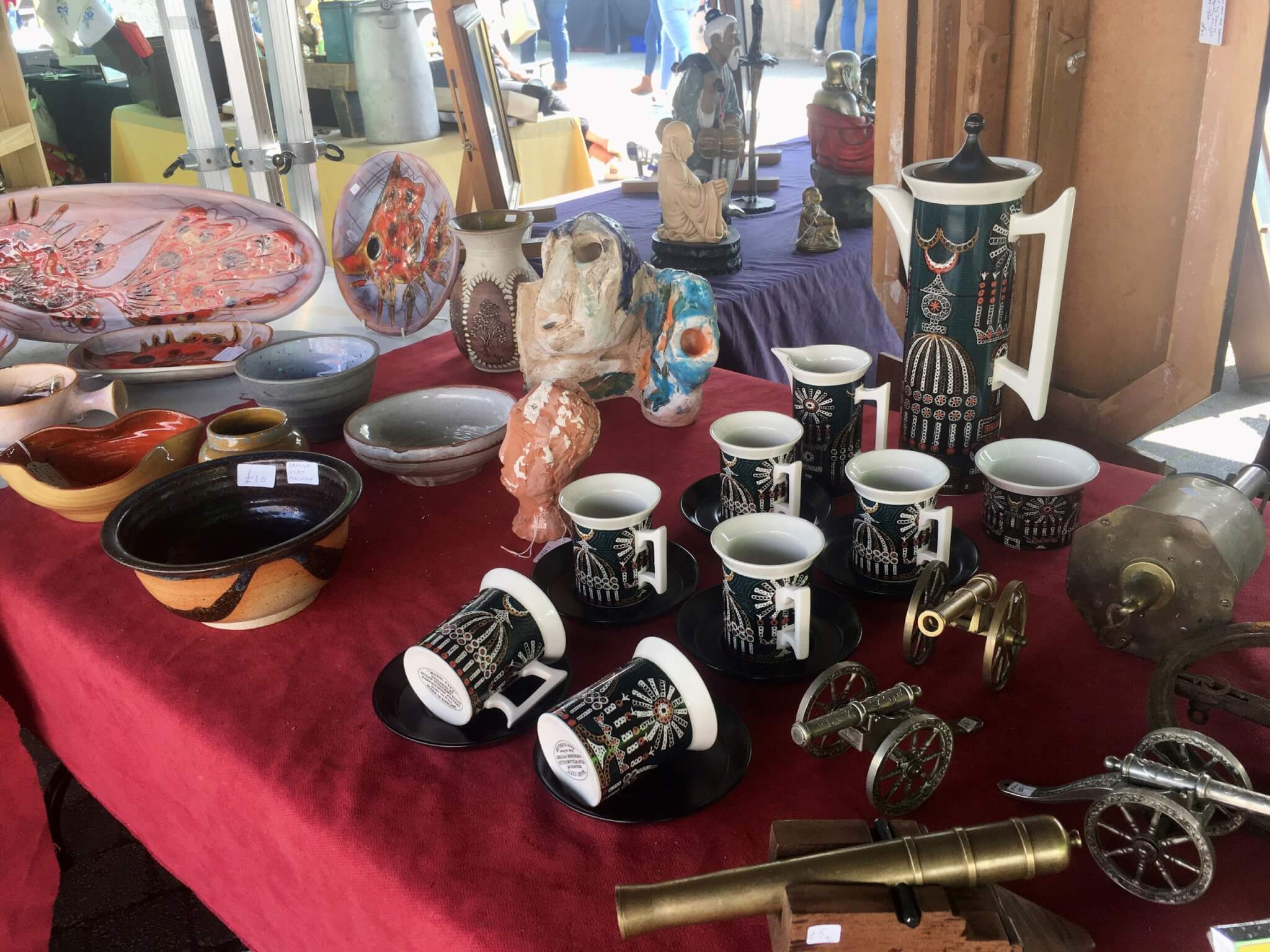 Retro coffee set, studio pottery and brass cannon ornaments on a red-covered antiques stall in Truro