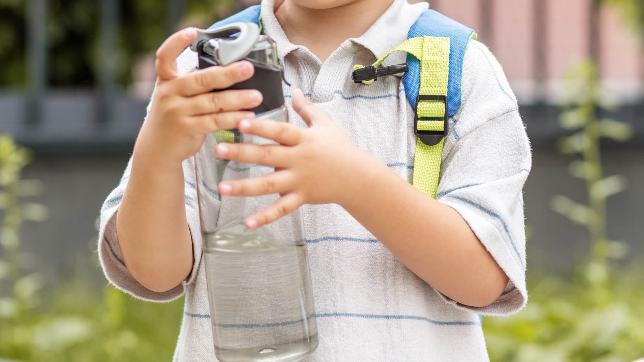 A child holding a water bottle while wearing a school backpack