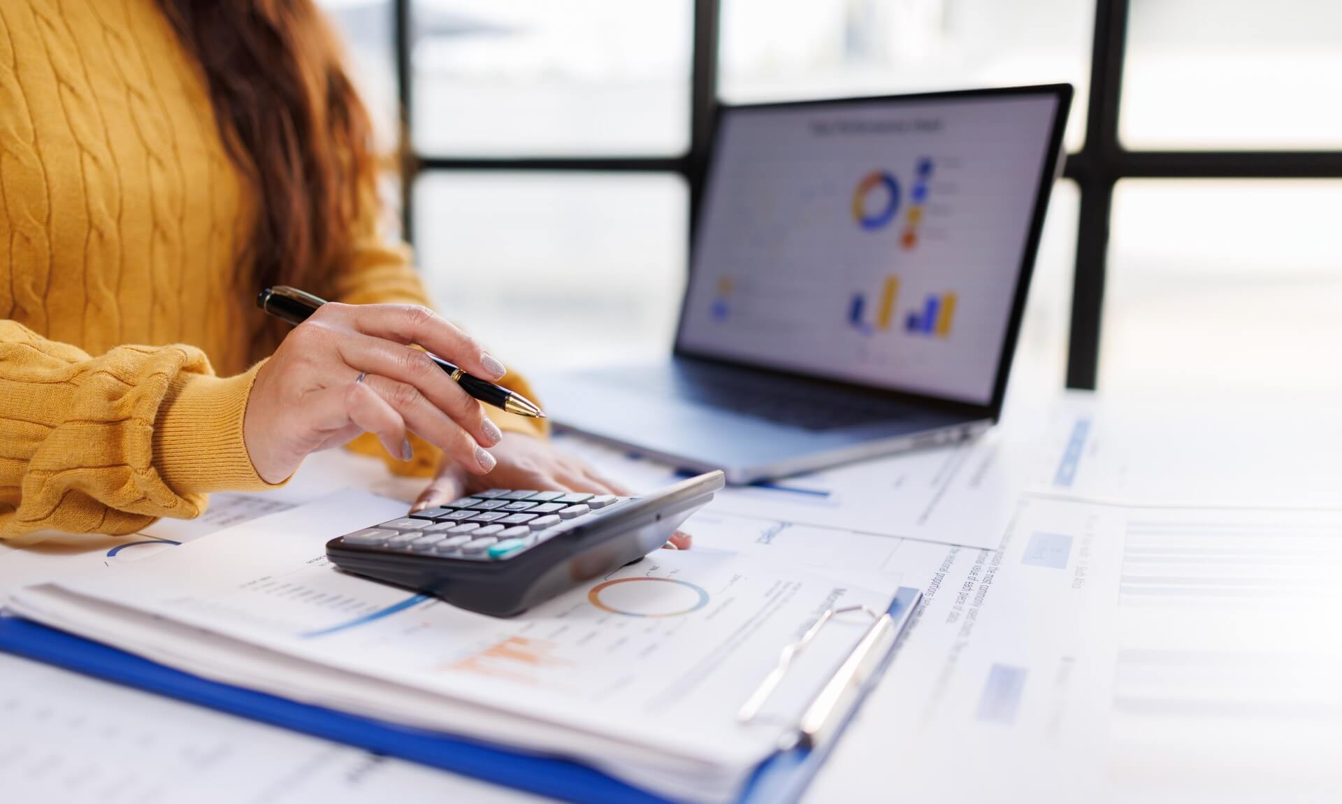An accountant working through business figures on a calculator, with charts on a laptop screen behind