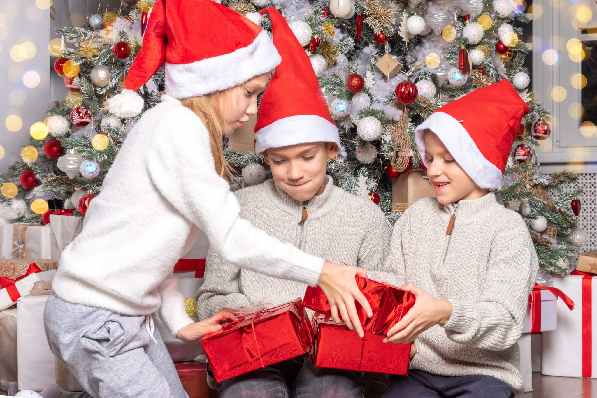 Three children in Santa hats opening red presents by a Christmas tree