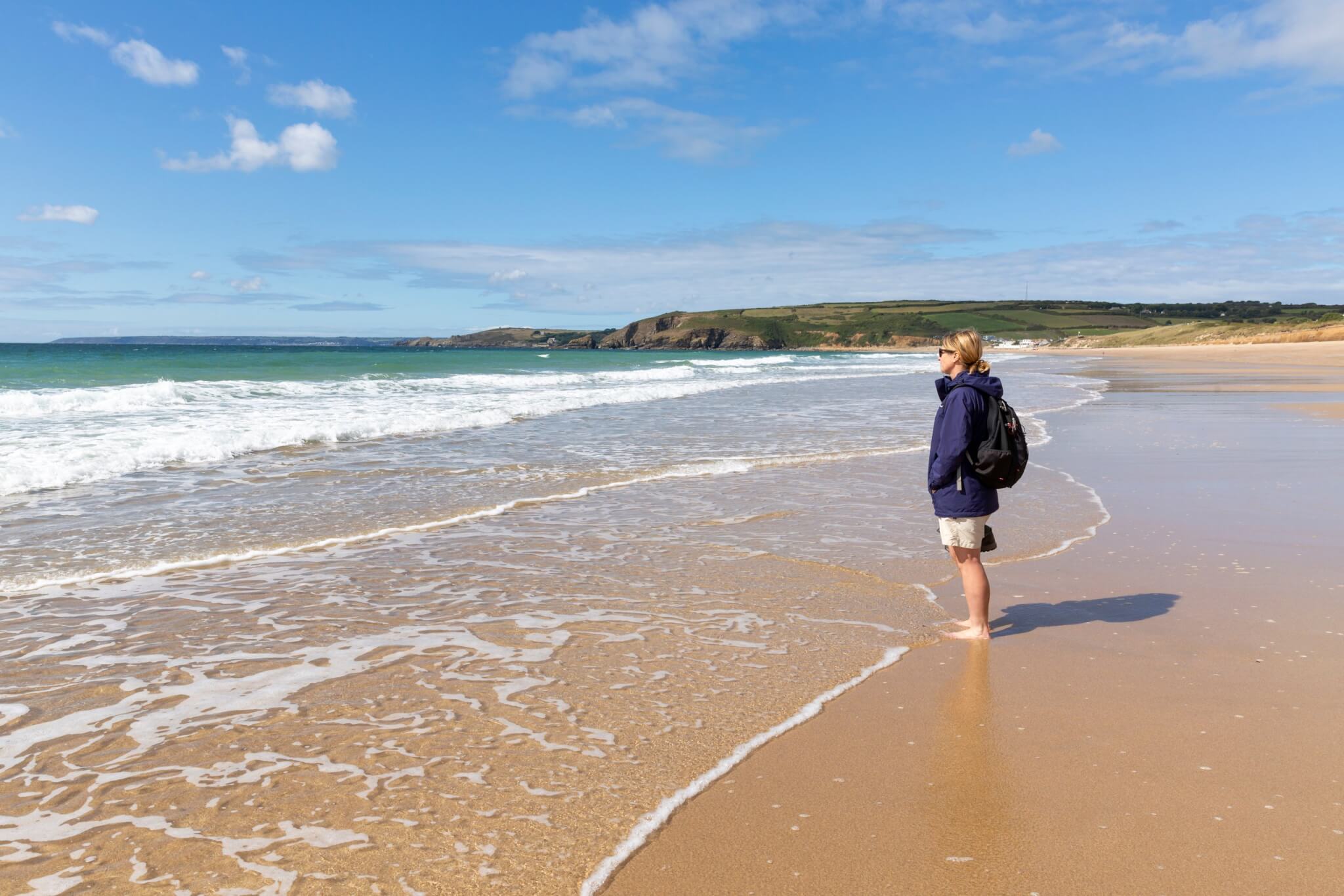 Woman standing barefoot at the water's edge on a Cornish beach looking out to sea