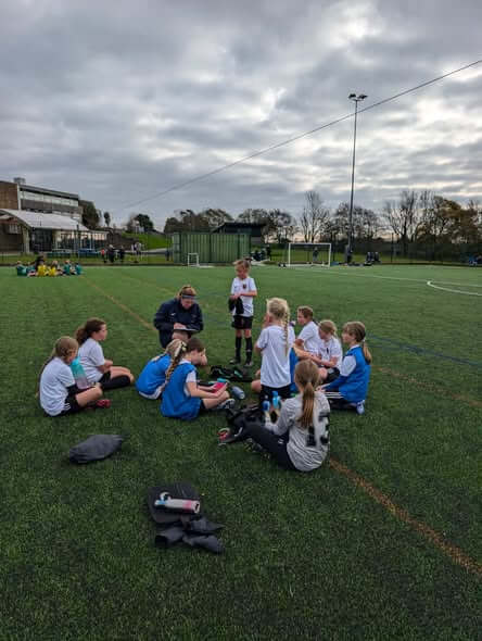 A coach talking to a group of seated young footballers on a training pitch