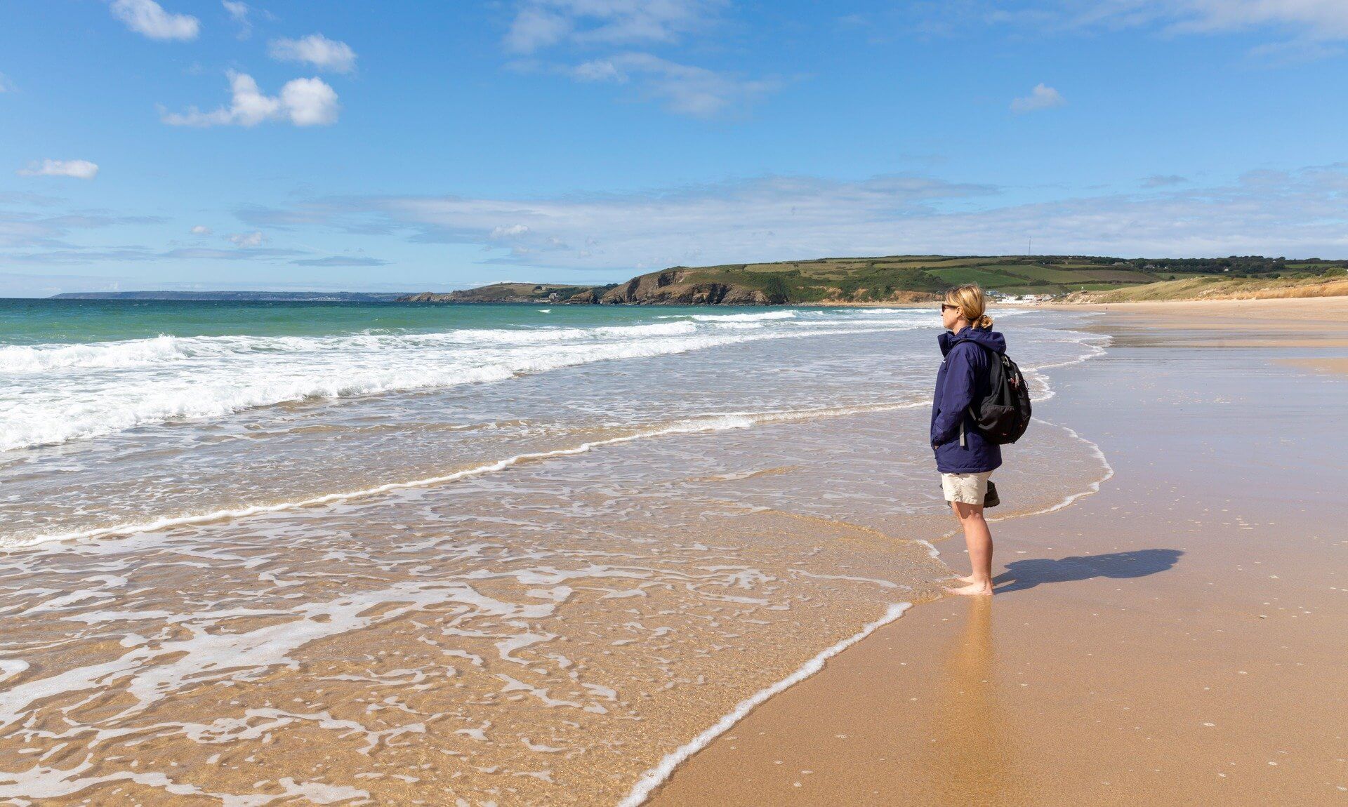 Woman standing barefoot at the water's edge on a Cornish beach looking out to sea