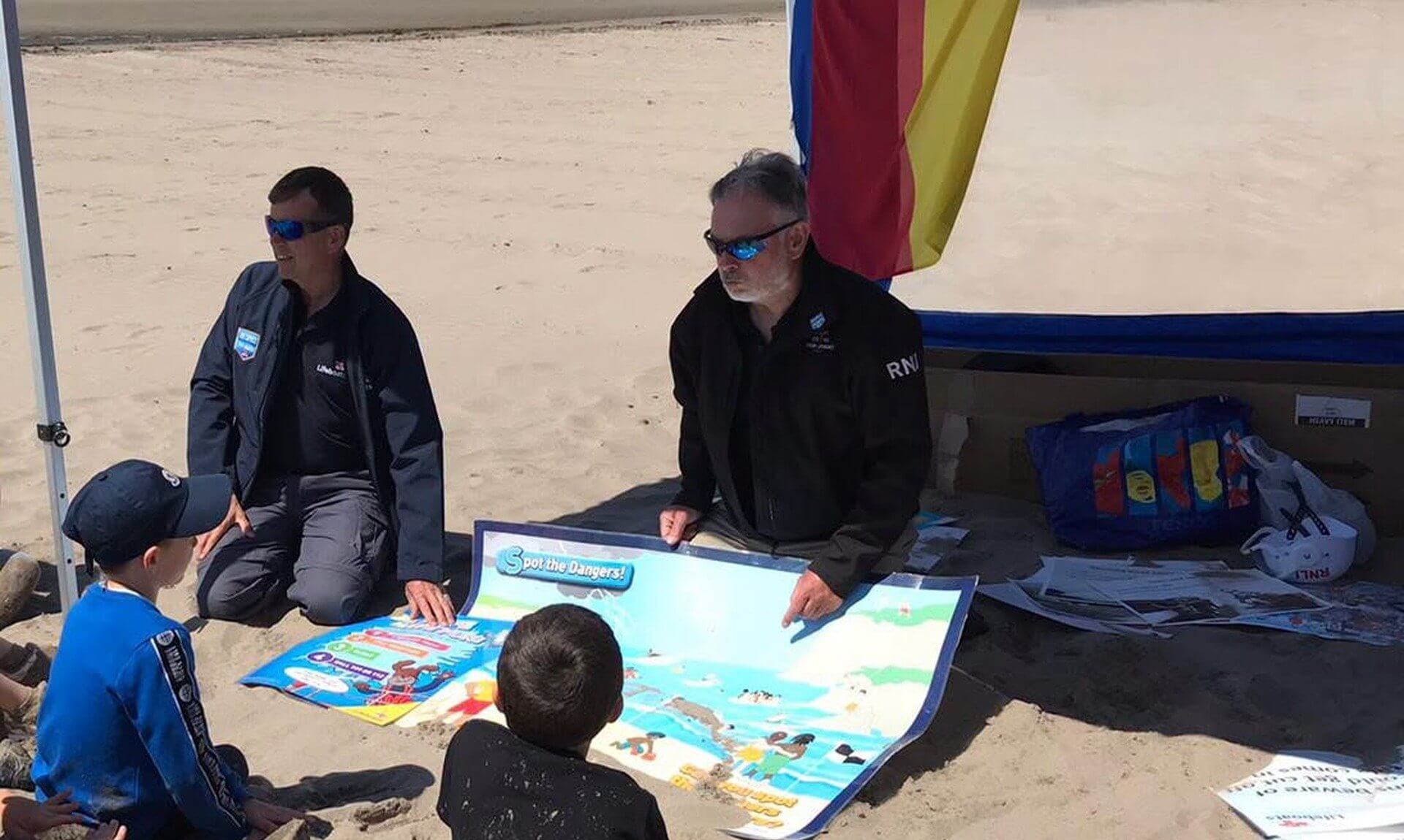 RNLI volunteers showing children a Spot the Dangers water safety poster on a beach