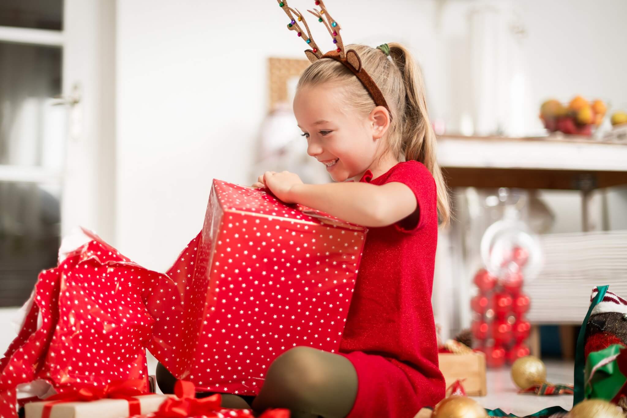 Young girl in reindeer antlers opening a red Christmas present