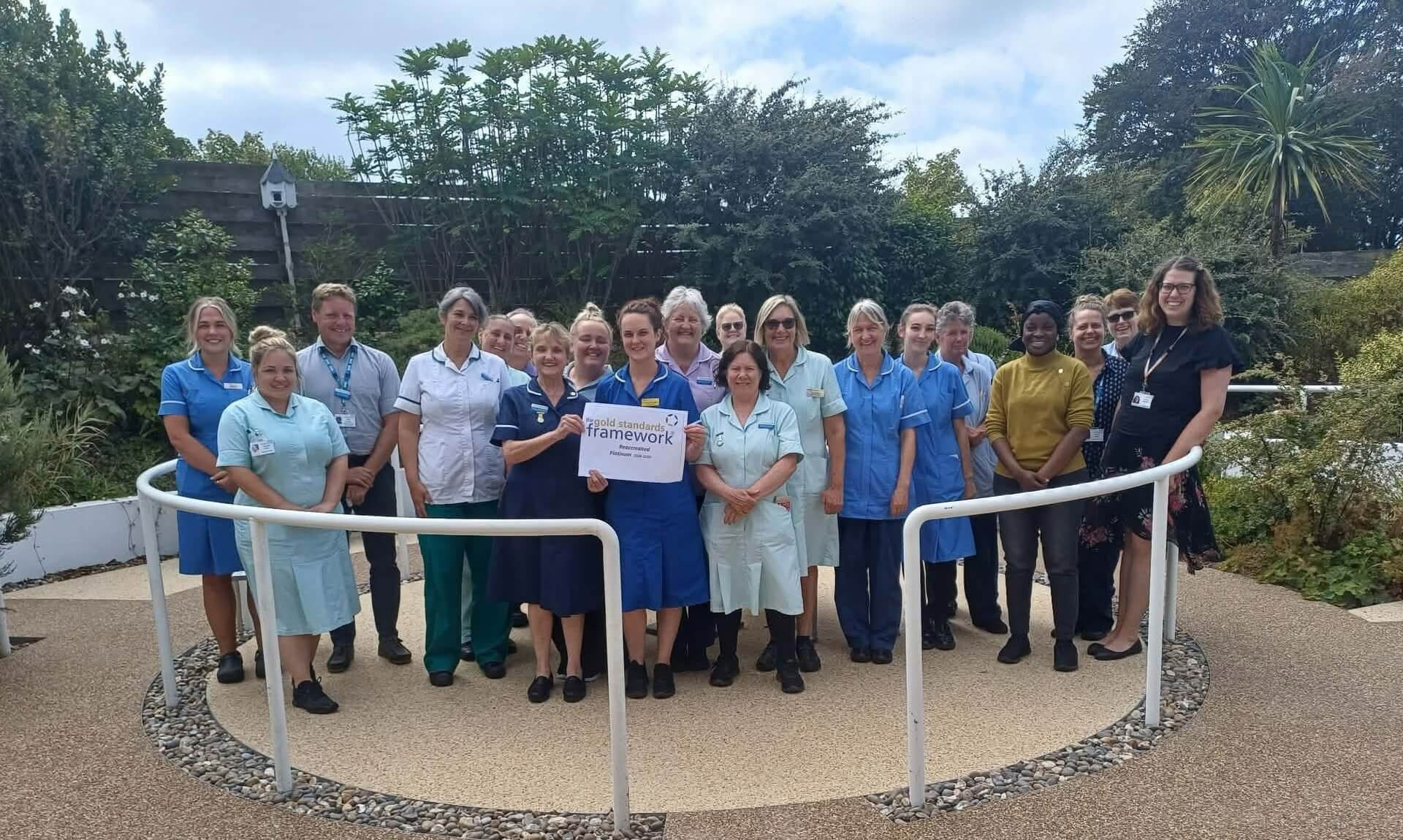 Helston Community Hospital staff holding a Gold Standards Framework platinum reaccreditation sign in the hospital garden