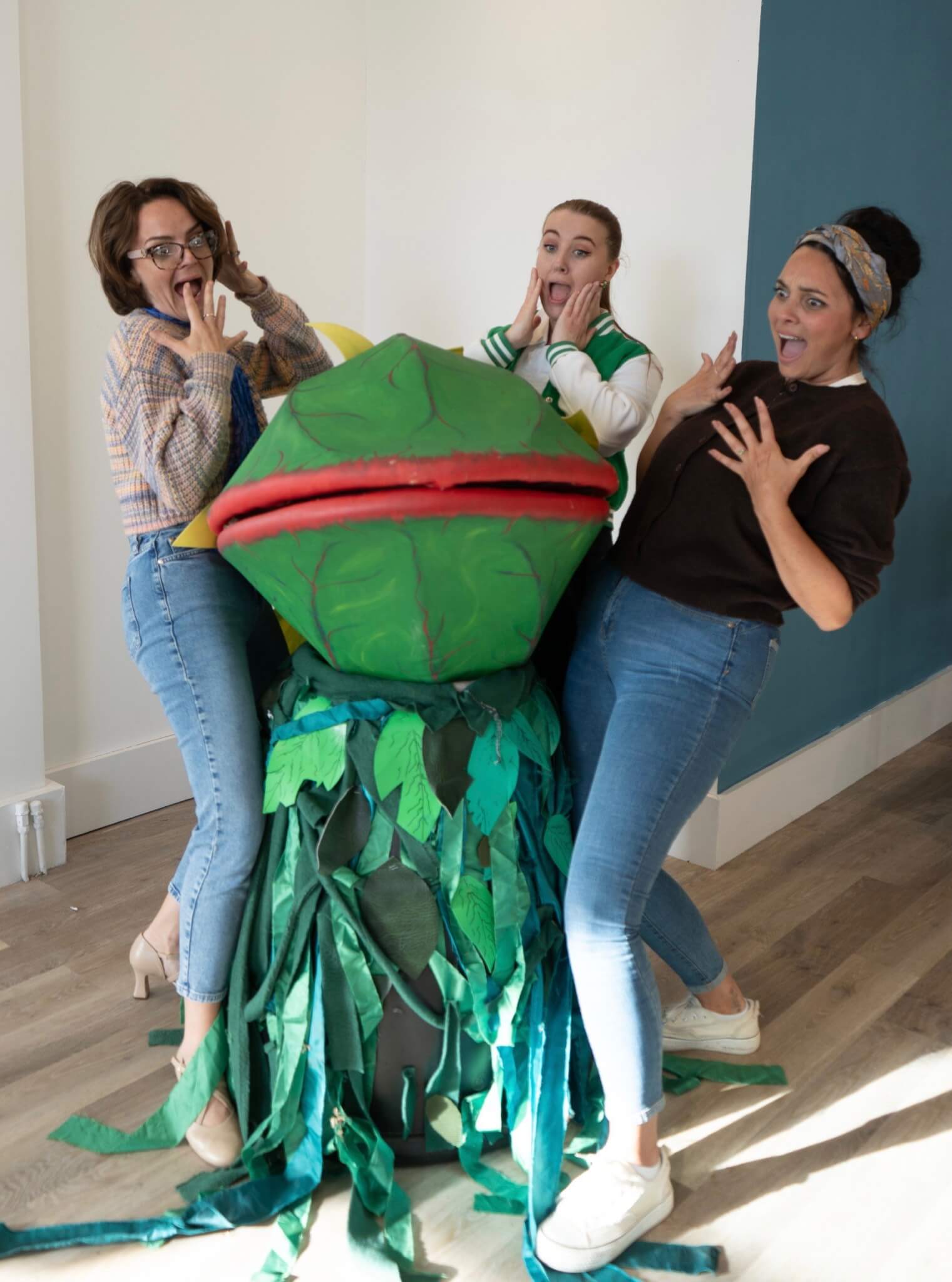 Three cast members pull shocked faces beside the Audrey II plant puppet