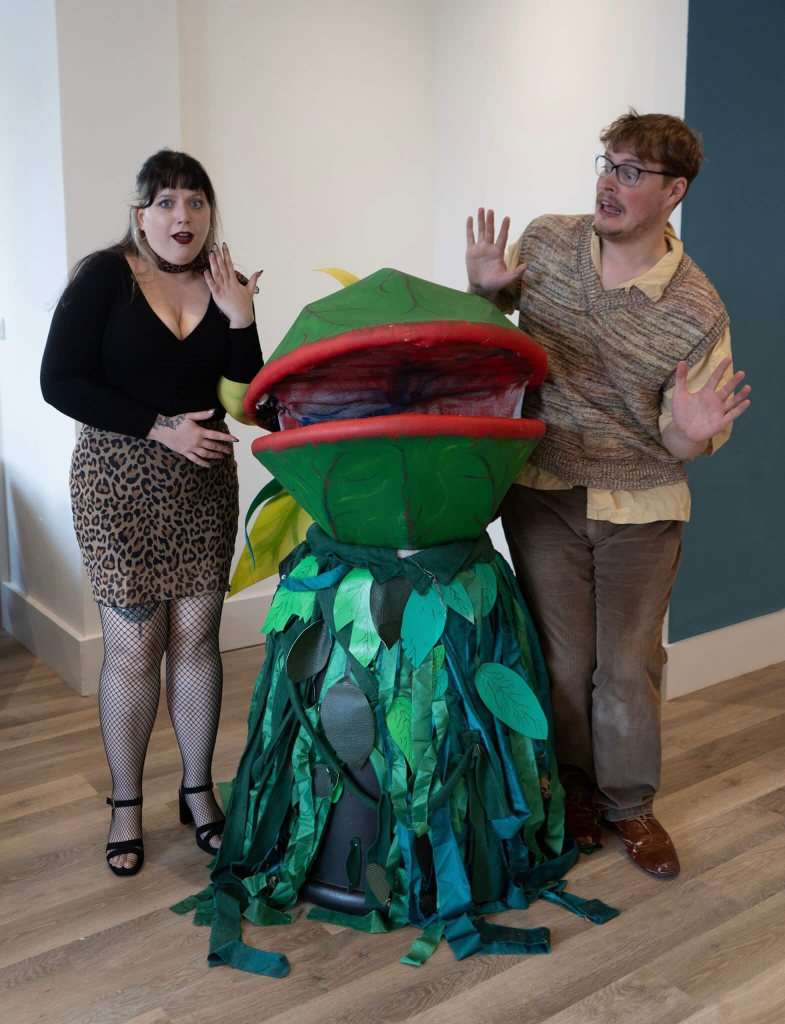 Two cast members look alarmed either side of the Audrey II plant puppet