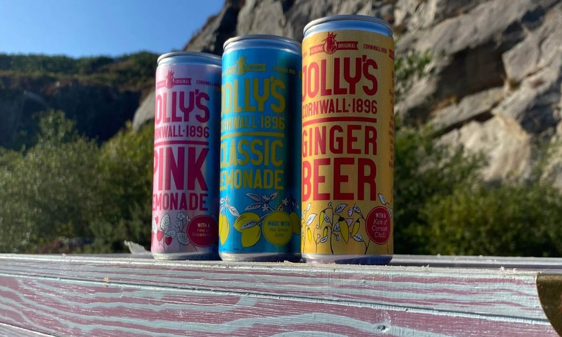 Cans of Jolly's Pink Lemonade, Classic Lemonade and Ginger Beer on a painted wooden set in front of a rocky cliff