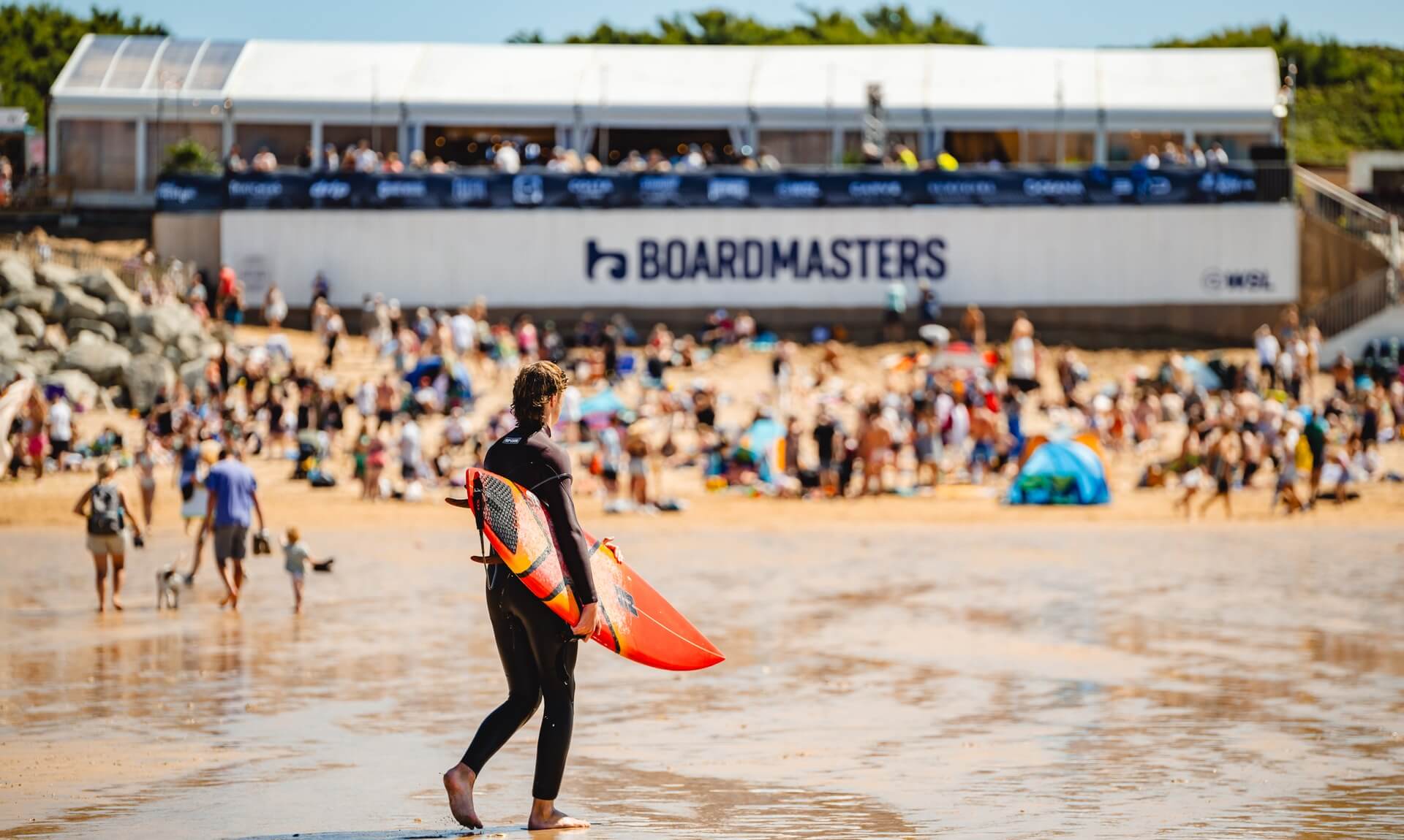A surfer with a board walking on the beach in front of the Boardmasters festival signage at Fistral