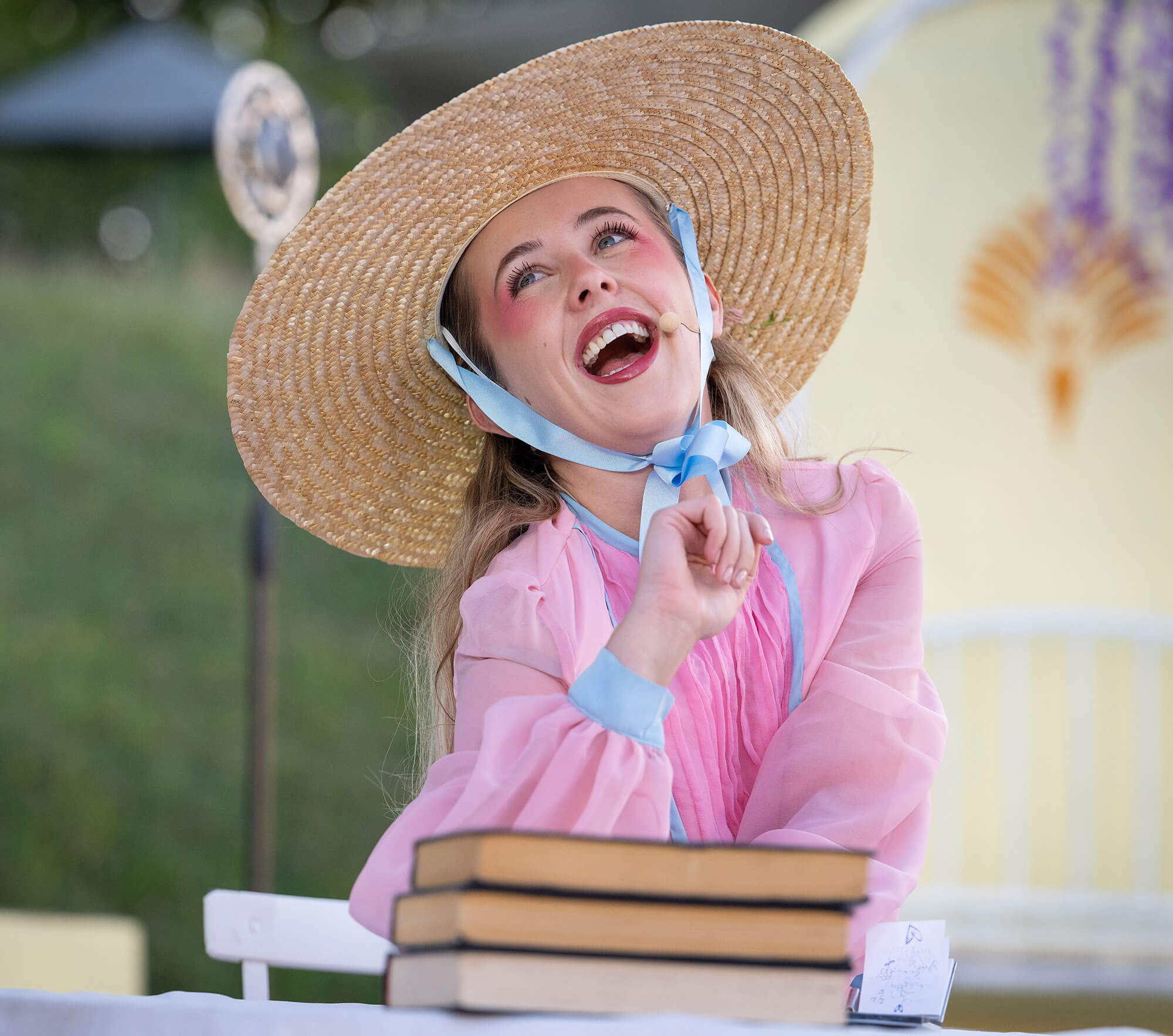 An actor in a straw hat laughing with a stack of books