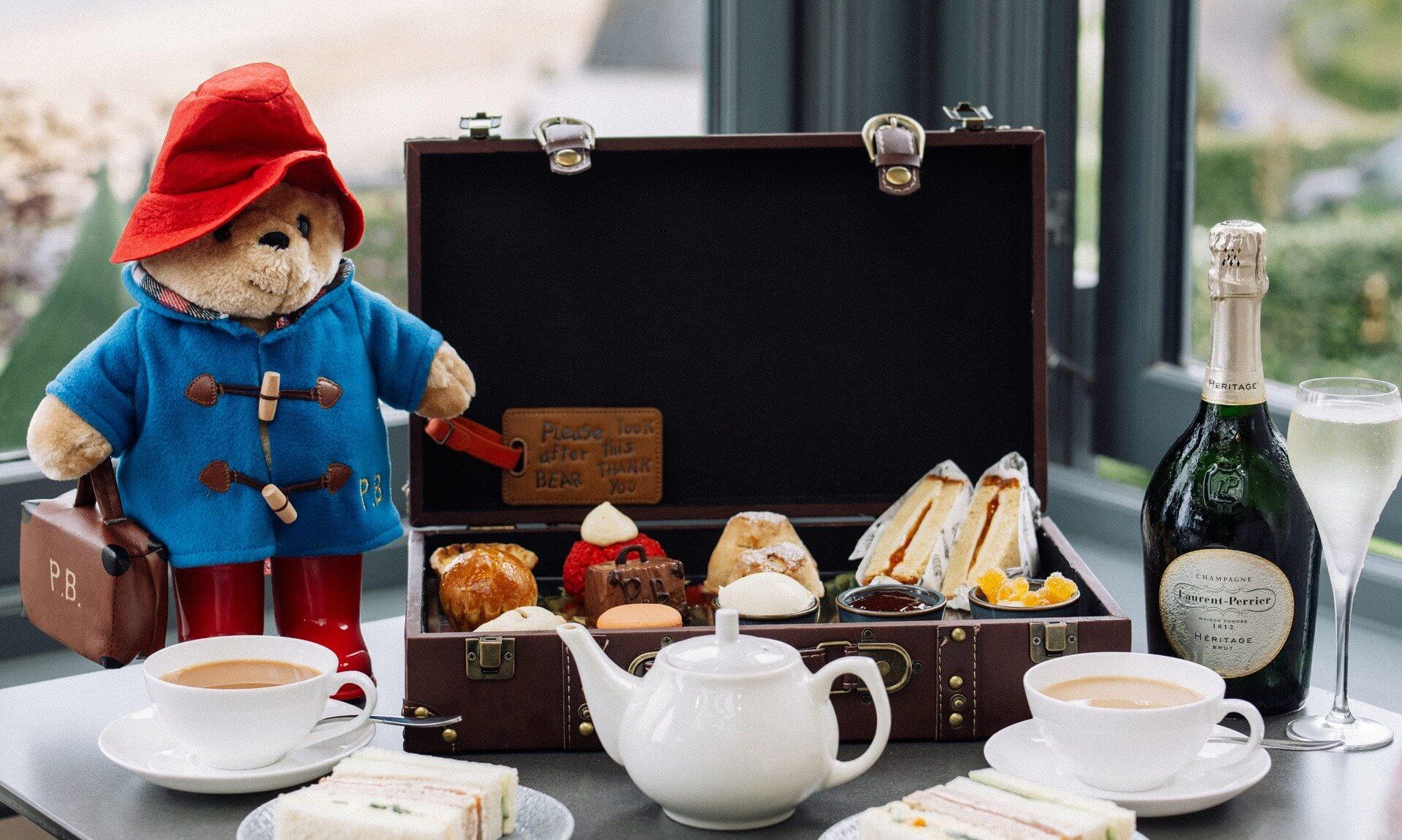 Paddington Bear beside an open suitcase of afternoon tea treats, with tea, sandwiches and Champagne by the window at Carbis Bay