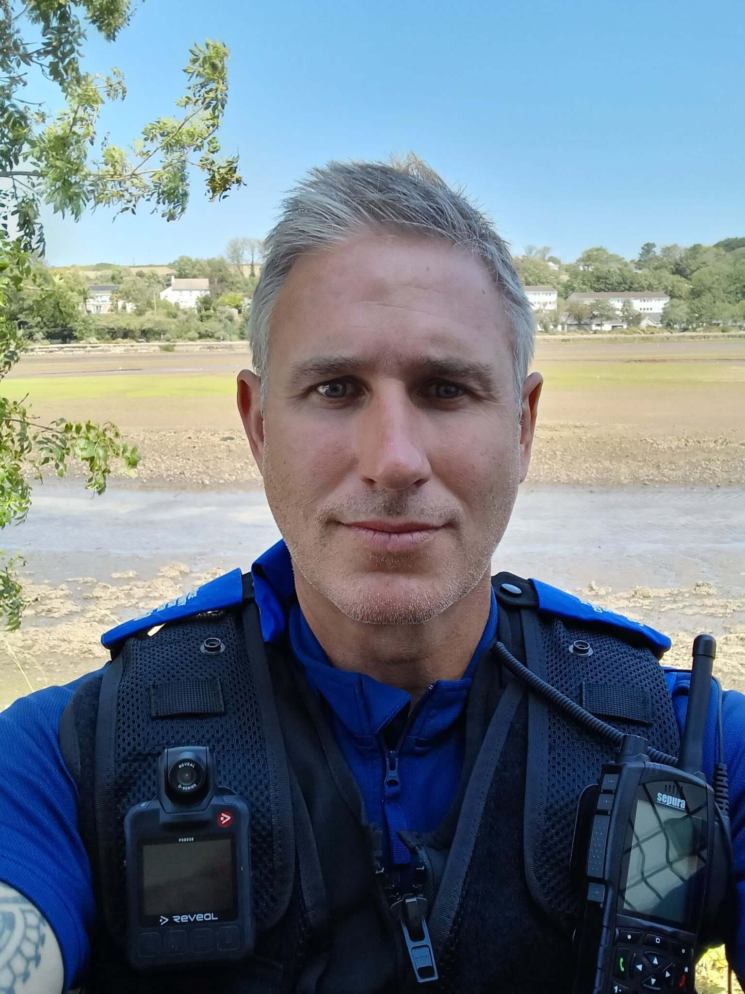 PCSO Aidan Jepson in uniform with mudflats and houses behind him