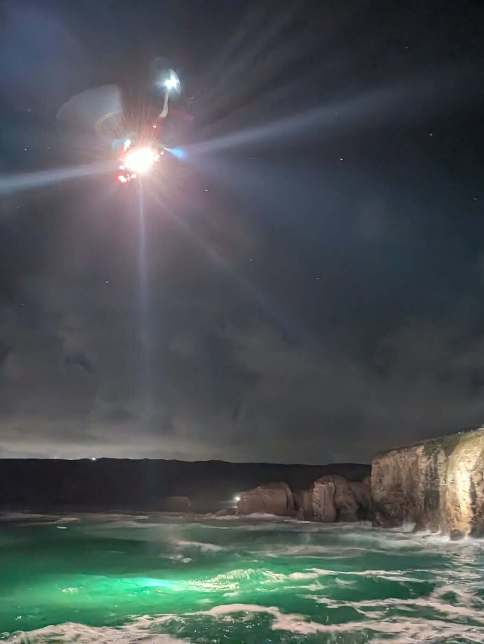 A rescuer's headtorch beam cuts through the night sky above the cliffs and sea at Perranporth