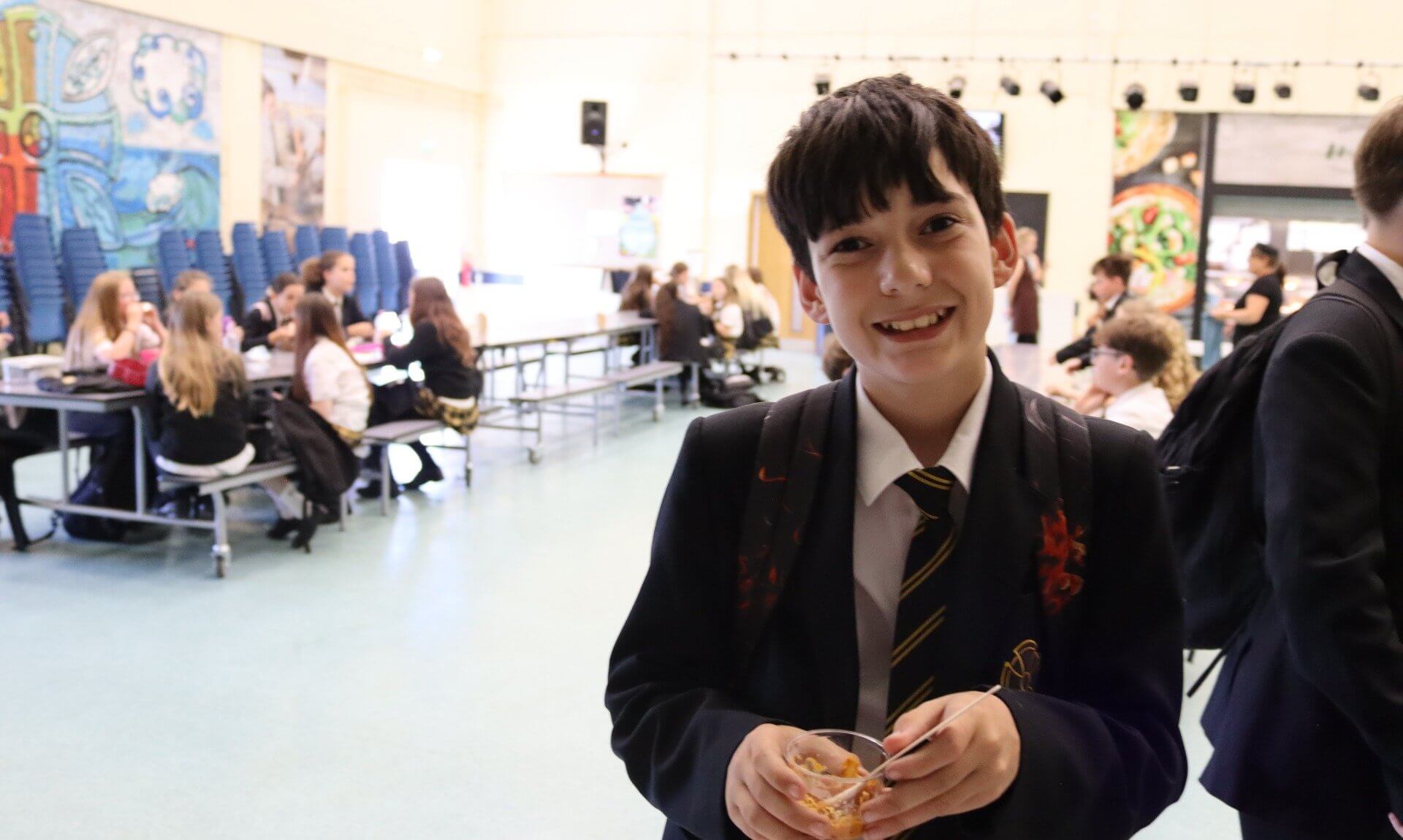 A smiling Pool Academy student holding a pot of food in the school hall