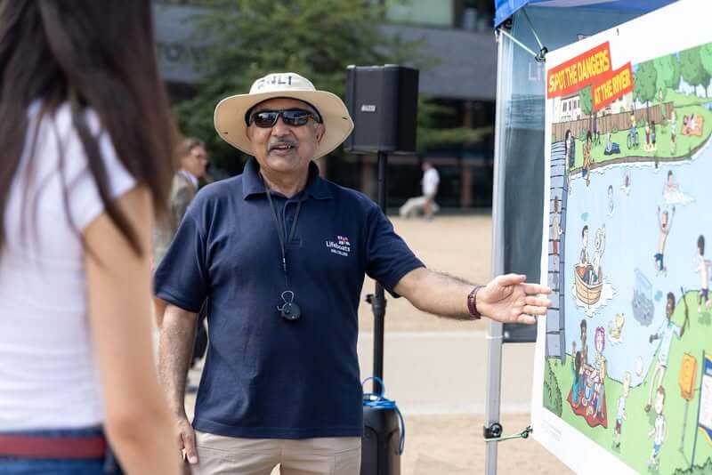 RNLI volunteer at a water safety stand next to a poster about spotting river dangers