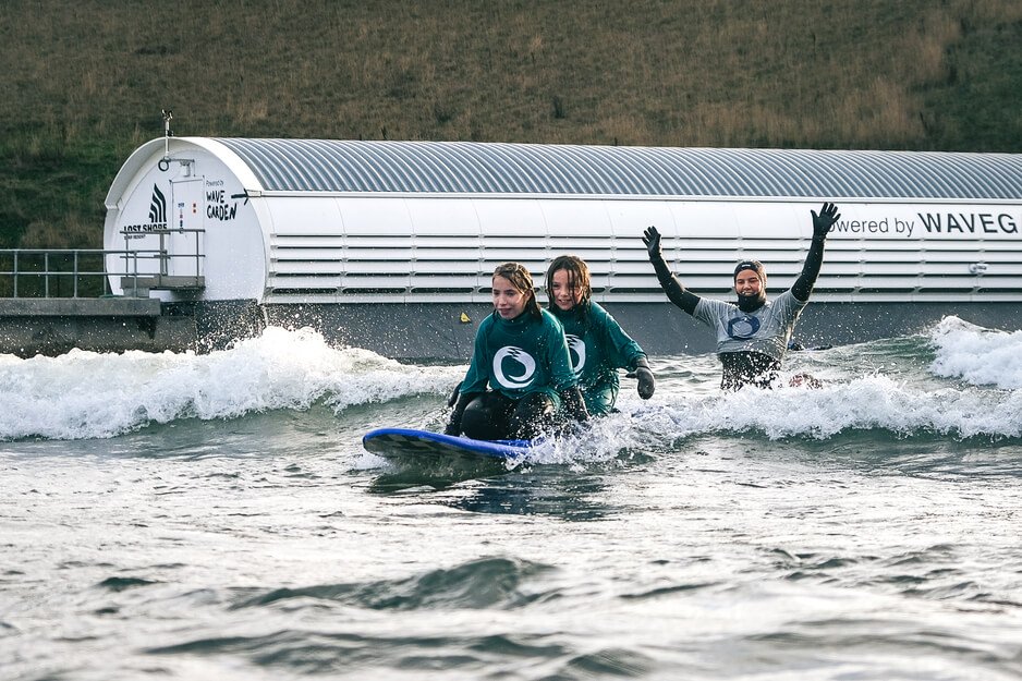 Two young surfers riding a board together at Lost Shore Surf Resort as a mentor cheers