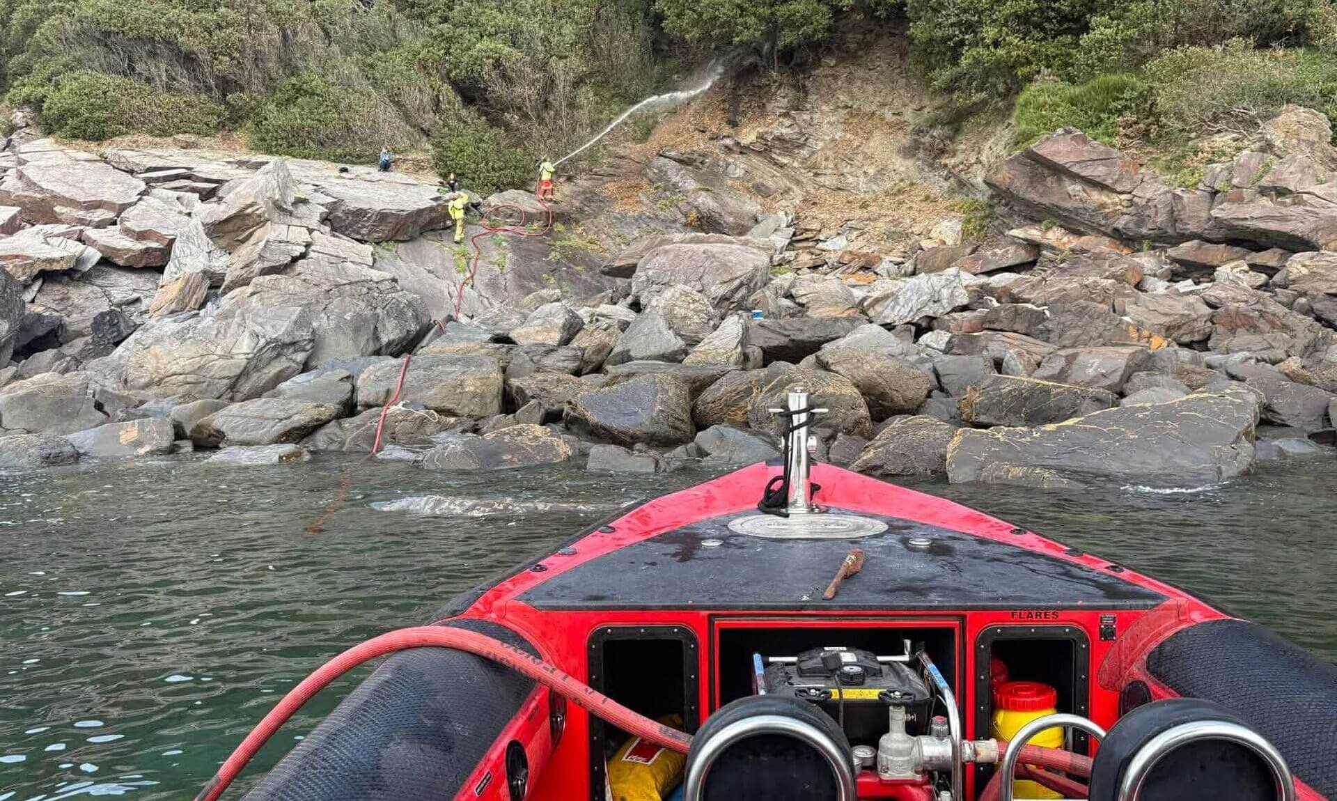 Falmouth fire boat off the rocky shore as firefighters spray water onto a wooded slope near Mawnan