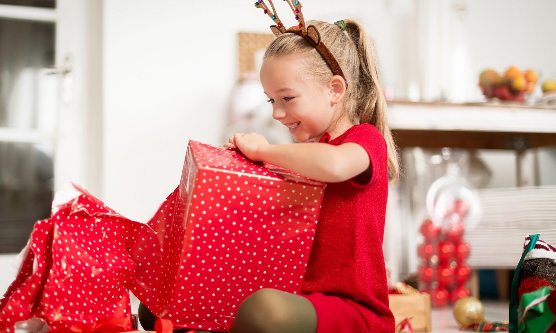 Young girl in reindeer antlers opening a red Christmas present