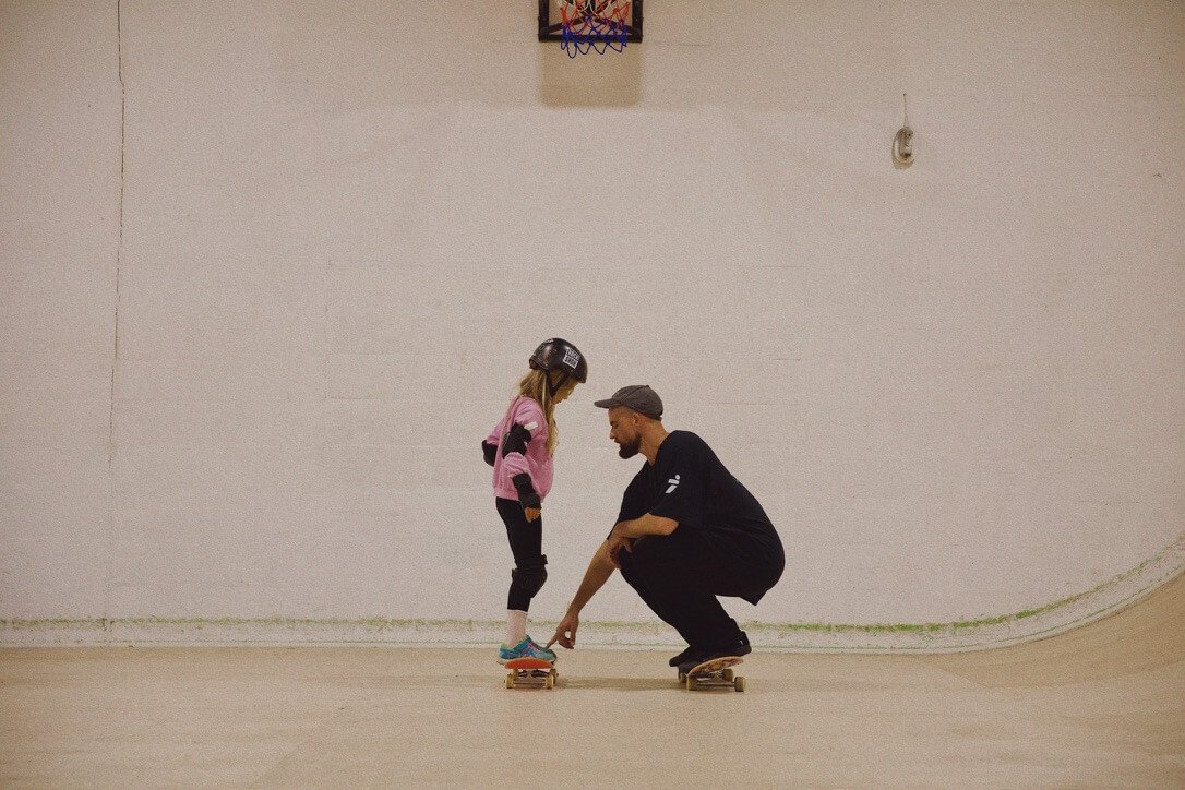 A TR7 coach crouching beside a young girl on a skateboard during a lesson