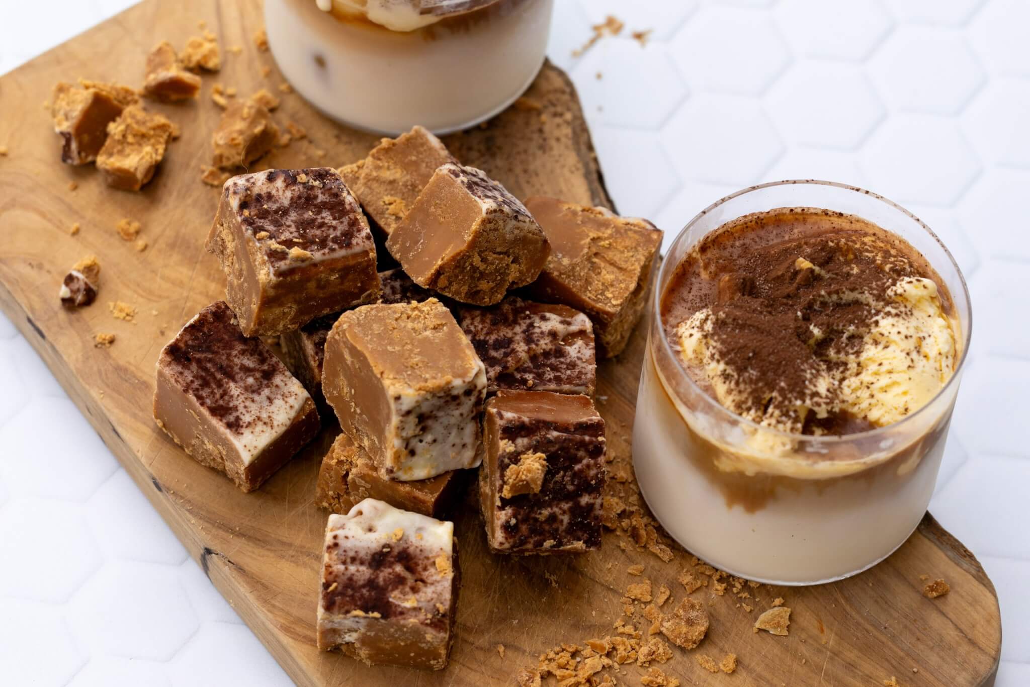 Roly's Tiramisu Fudge pieces on a wooden board beside a glass of tiramisu