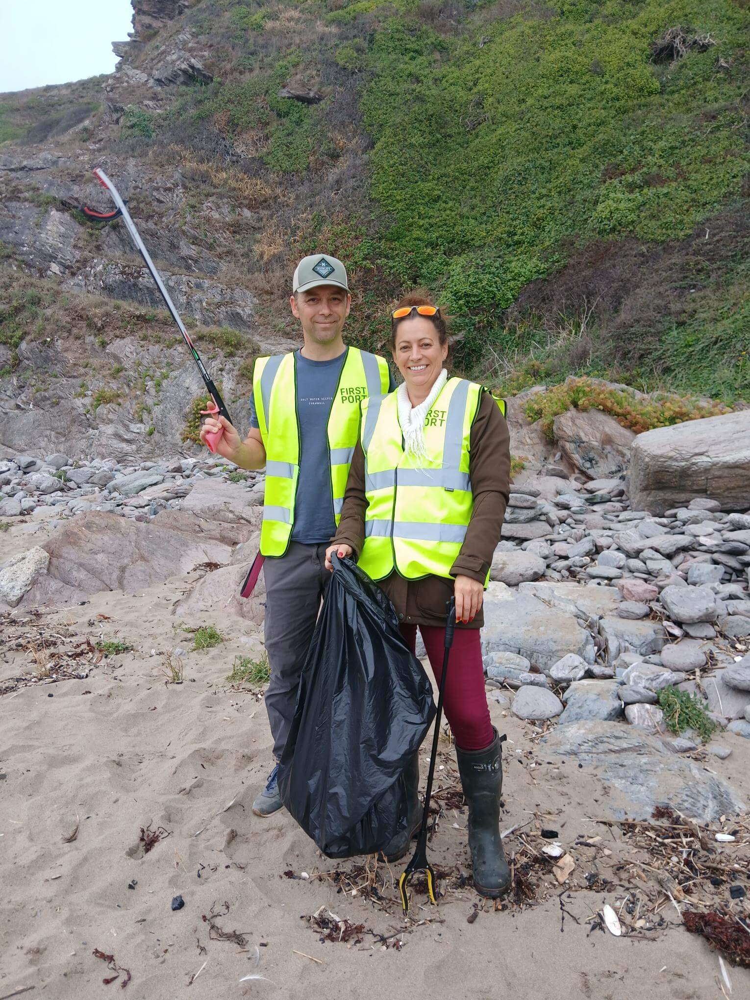 Two volunteers in FirstPort hi-vis vests holding a bin bag and litter pickers at Tregonhawke Beach