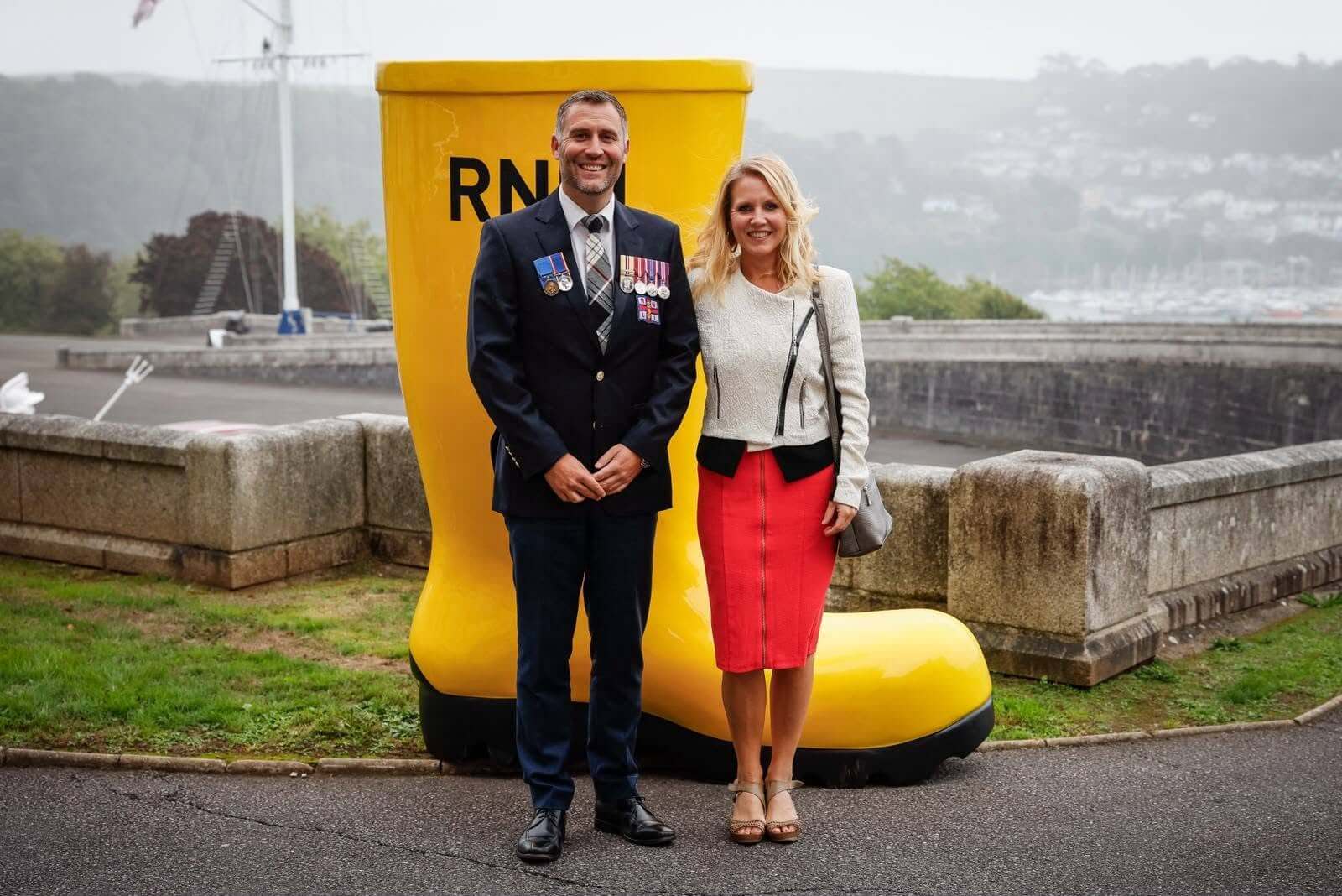 A man wearing medals and a woman standing in front of a giant yellow RNLI welly at Britannia Royal Naval College