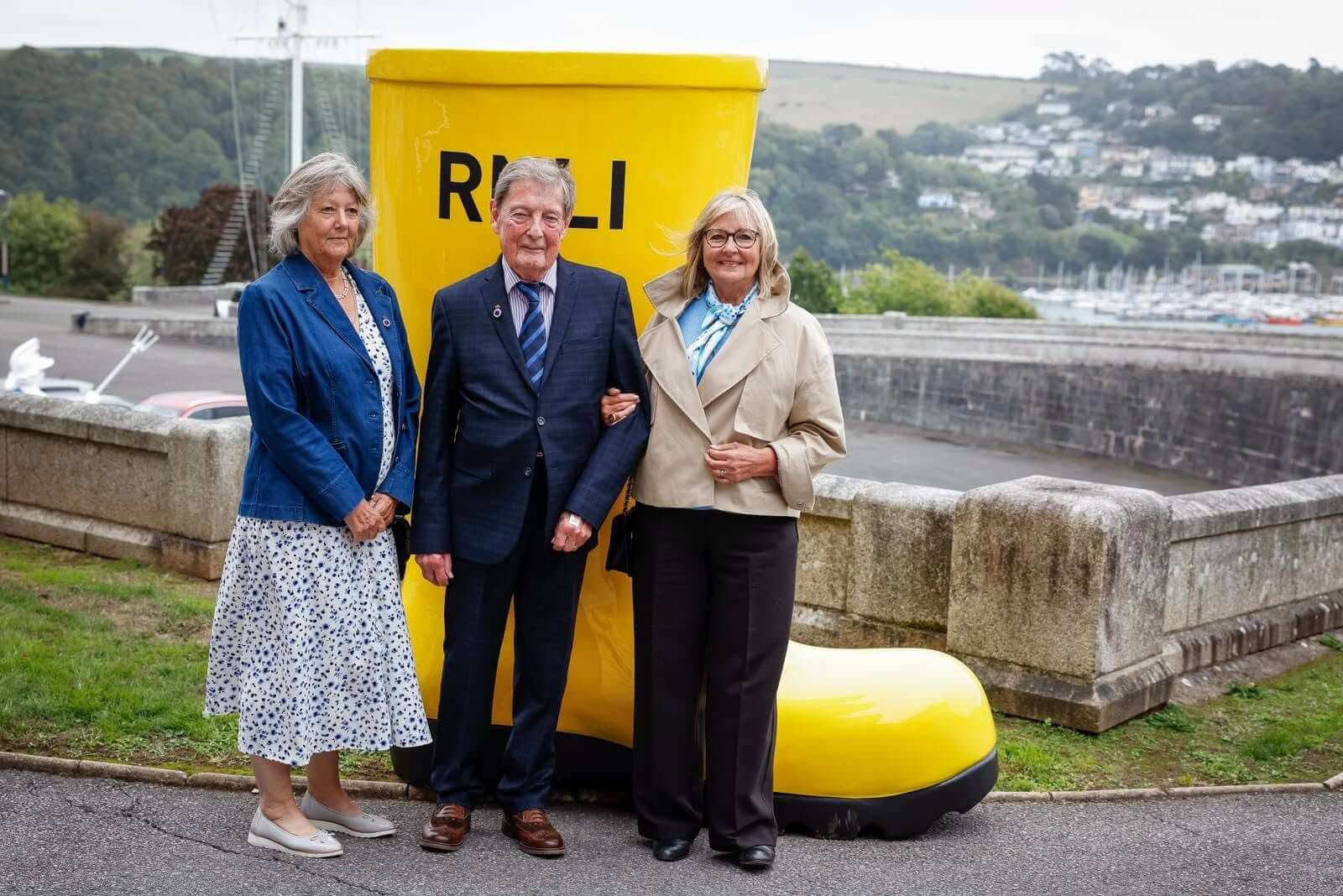 Three people standing in front of a giant yellow RNLI welly at Britannia Royal Naval College