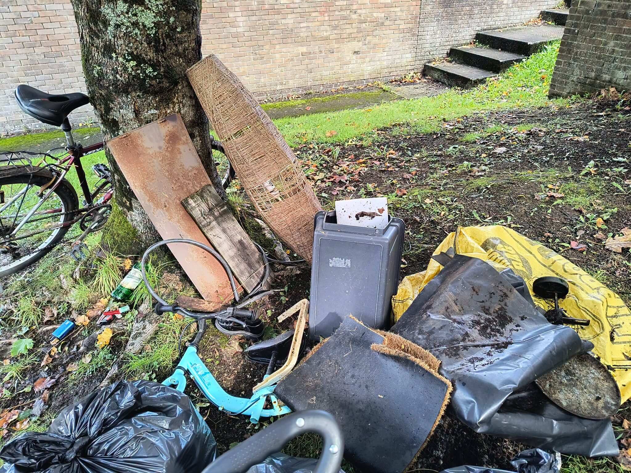 Fly-tipped bikes, lamp shade, doormat and other items piled by a tree in Bodmin