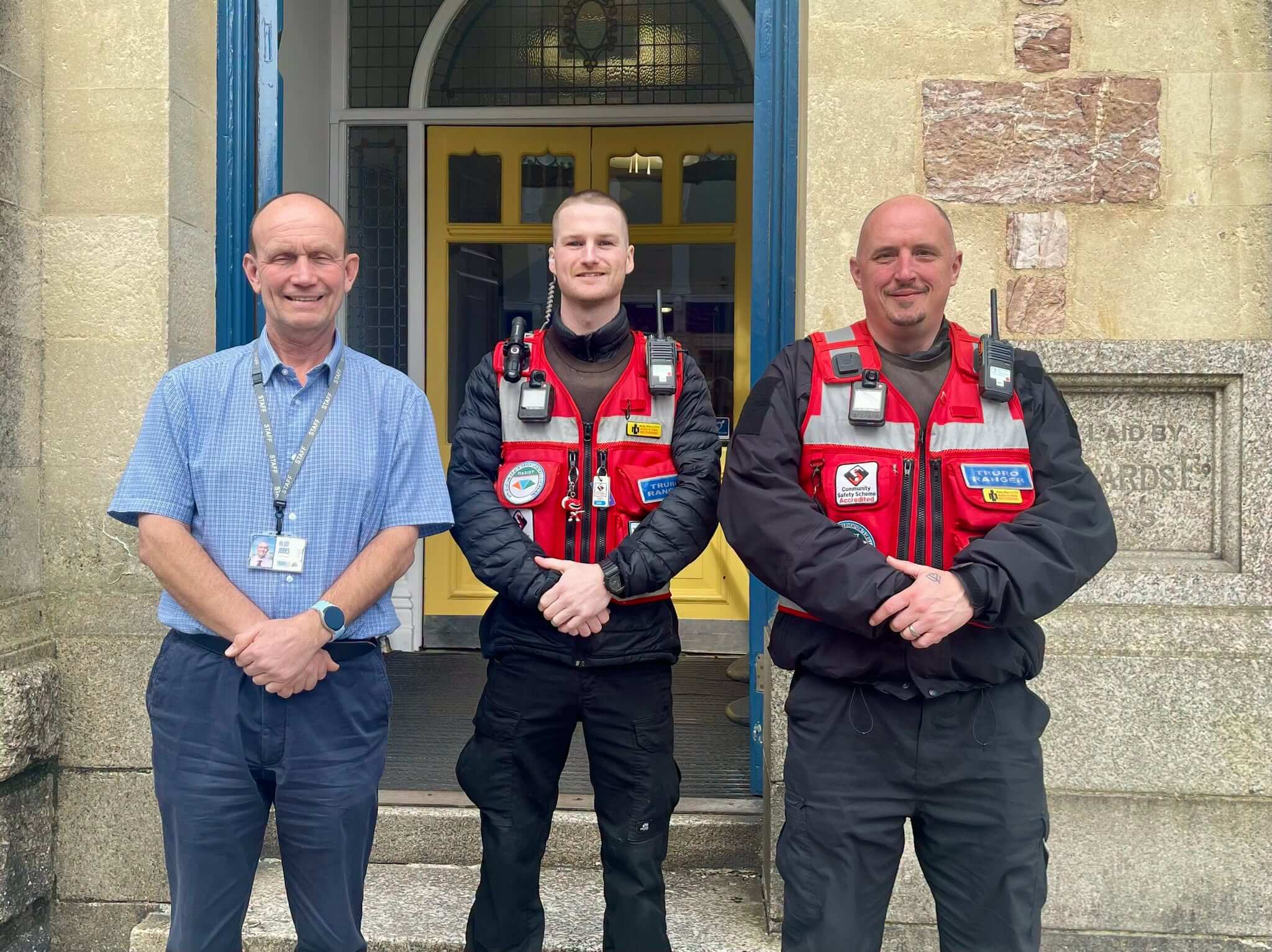 Two Truro Rangers in red patrol vests standing beside a man in a blue shirt outside a building in Truro