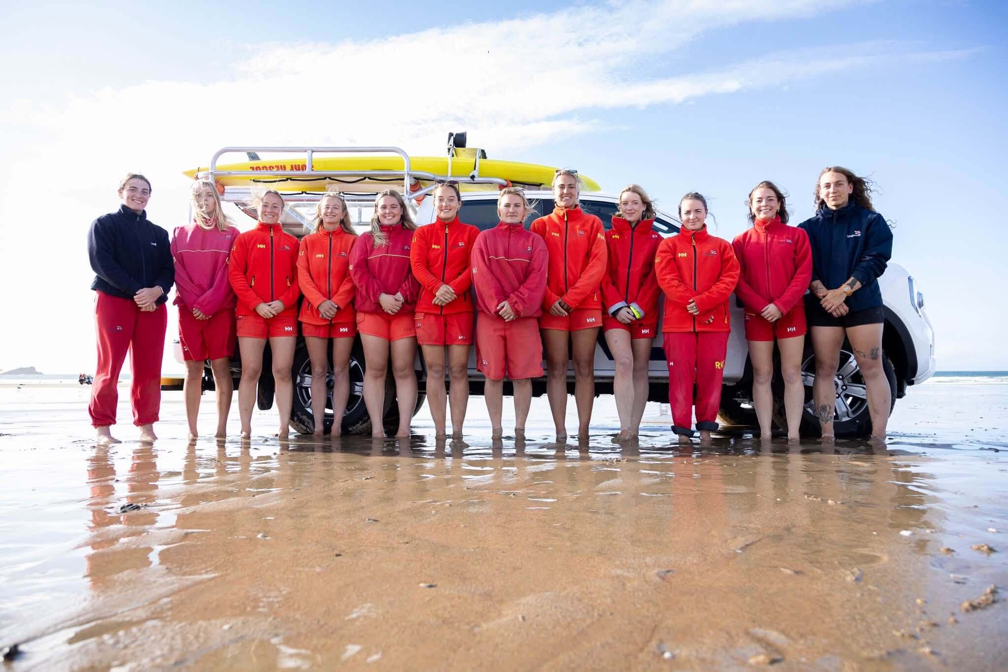 Twelve women in RNLI lifeguard uniform standing in front of a lifeguard truck on the beach