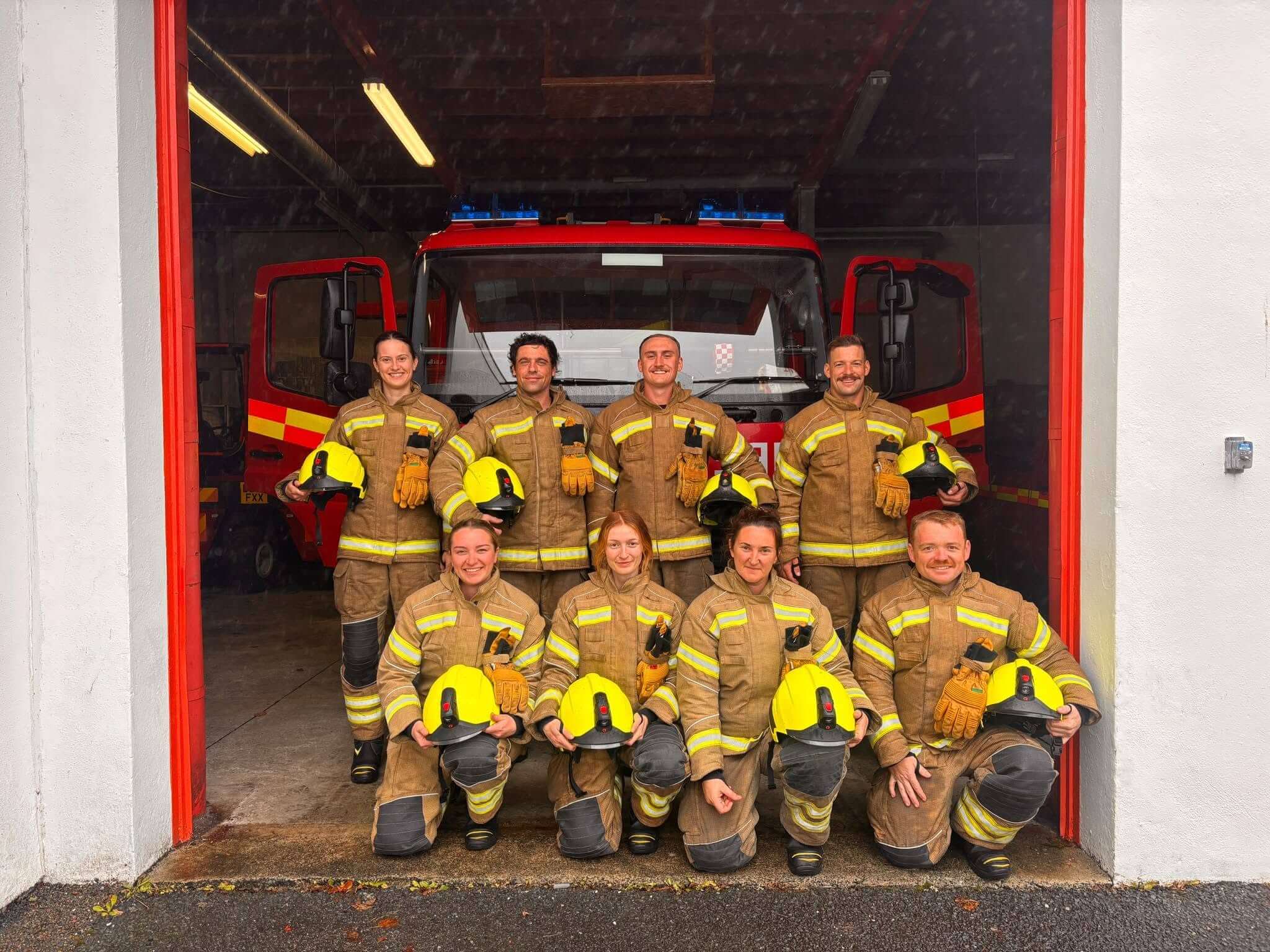 Eight firefighters in kit in front of a fire engine at a fire station