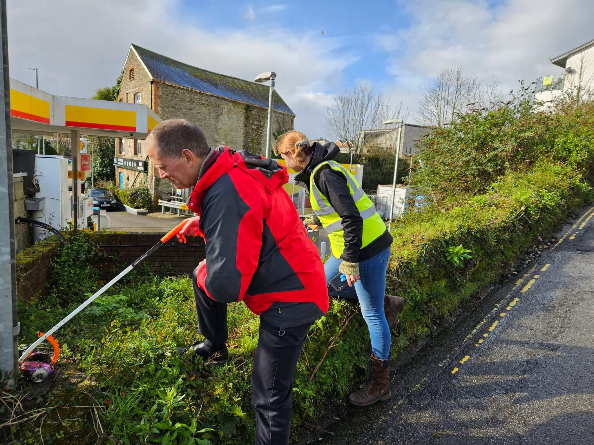 Two volunteers litter picking on a grass verge beside a petrol station