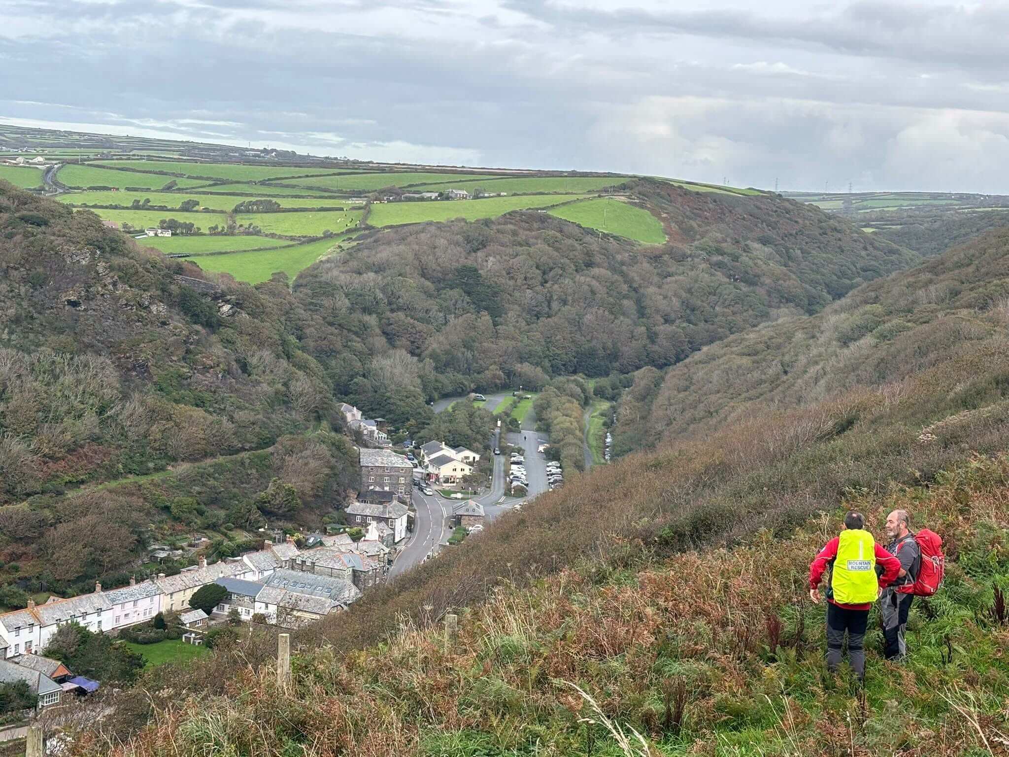 Two mountain rescue volunteers on a hillside looking down over Boscastle village and valley