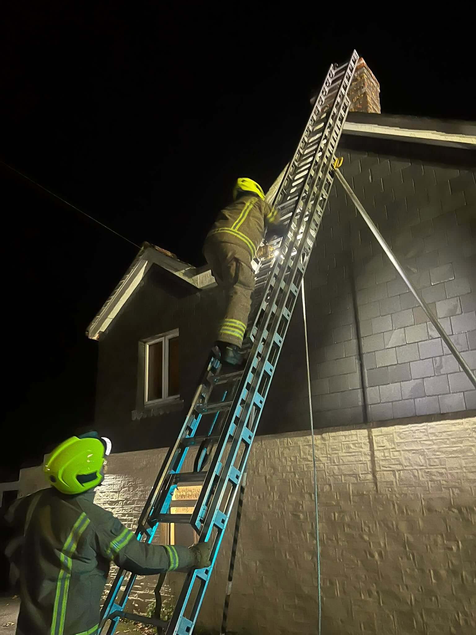 A firefighter climbing a ladder to a chimney at night while a colleague steadies it from below