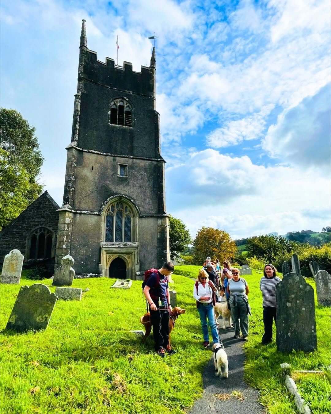 Walkers and dogs on a path through a churchyard below a church tower