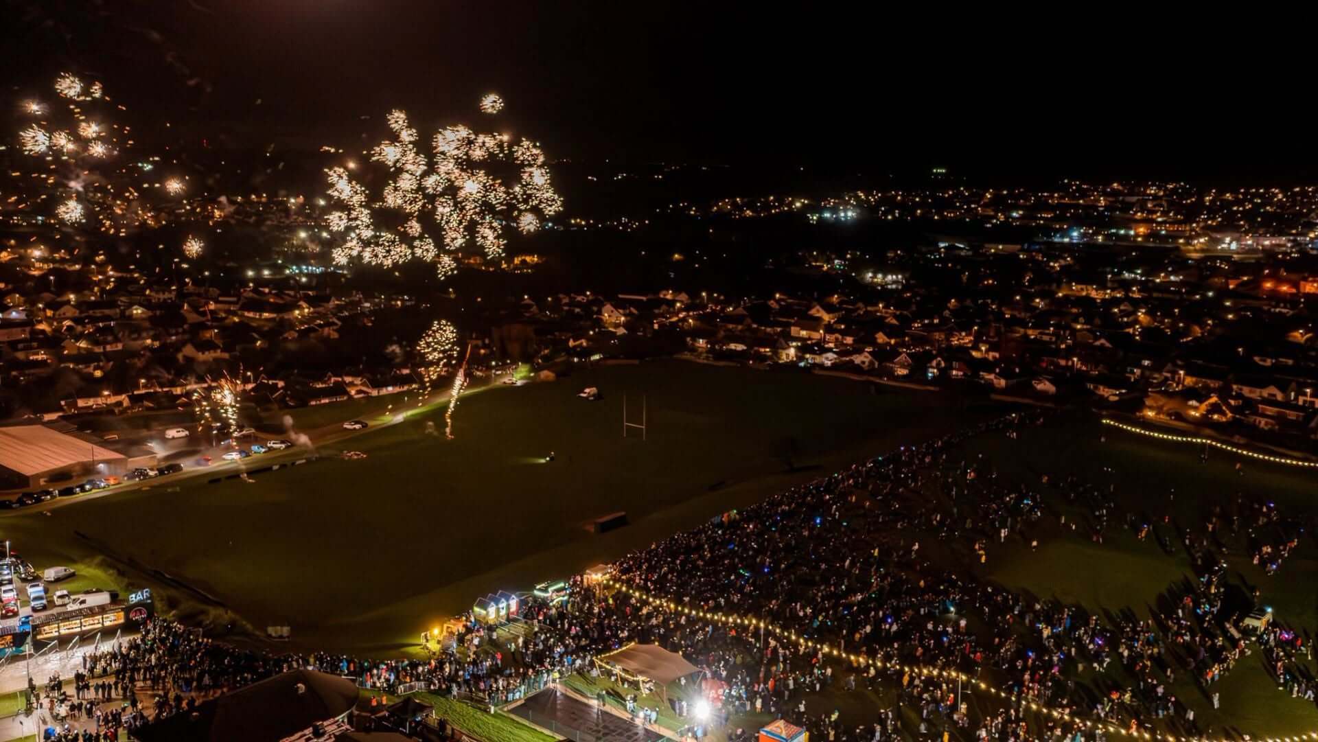 Fireworks over a town at night with crowds gathered on a playing field