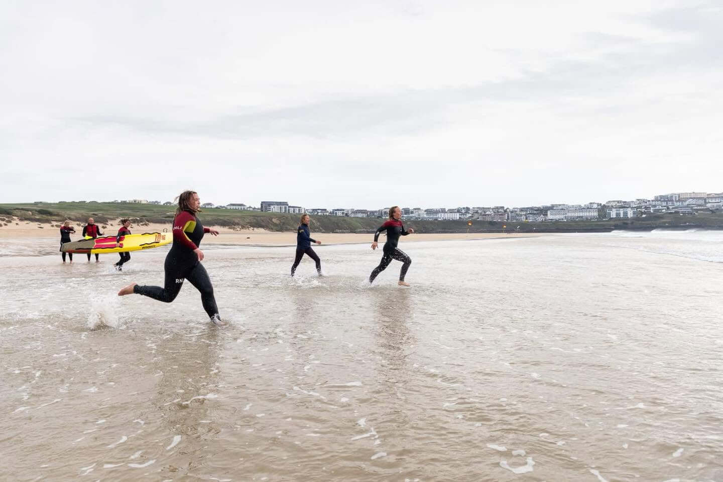 Lifeguards in RNLI wetsuits running through shallow water with a rescue board behind them