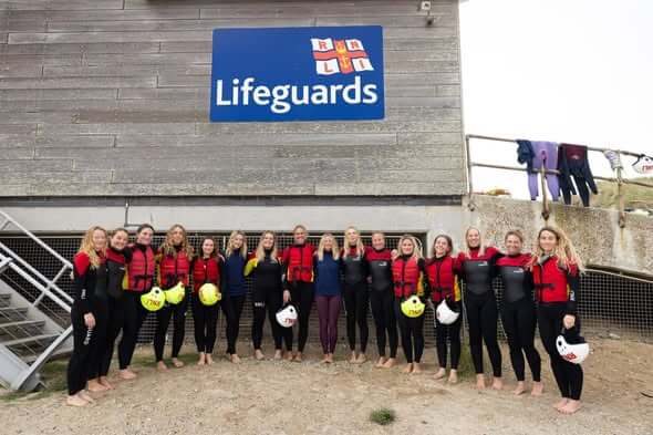 A group of lifeguards in wetsuits lined up beneath an RNLI Lifeguards sign
