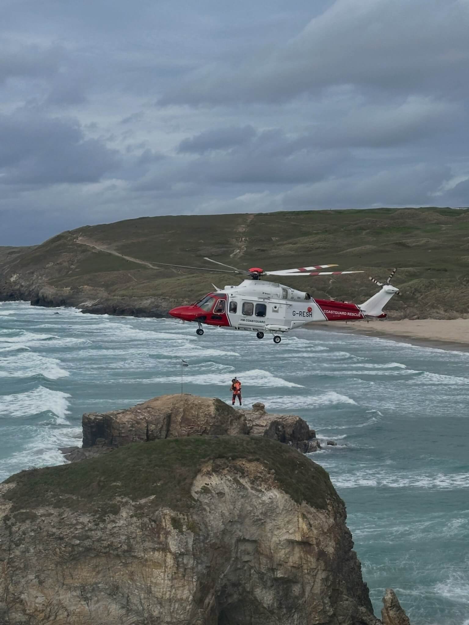 Coastguard rescue helicopter hovering over a rocky outcrop at Droskyn, Perranporth, with a winchman on the wire