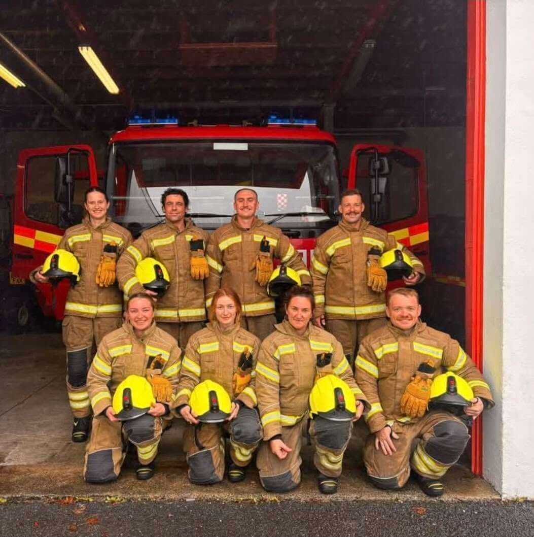 Eight firefighters in kit holding yellow helmets in front of a fire engine at Fowey Community Fire Station
